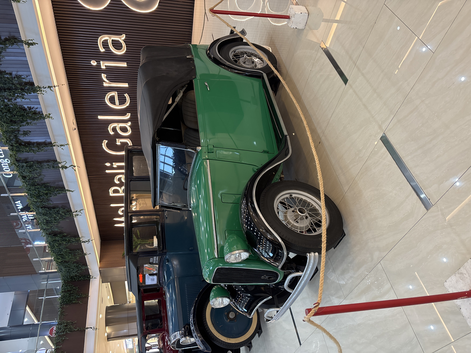 The image shows a display of vintage cars inside a shopping mall in Kuta, Indonesia. A green vintage convertible with a black top is the most prominent vehicle. In the background are other classic cars, including a dark blue sedan and a red car, all seemingly part of an exhibition or display. The mall’s interior is visible, with tile flooring and a wall featuring illuminated signage reading "Mal Bali Galeria". Potted plants are positioned on the upper levels of the mall. A rope barrier held by red poles is in place near the green car, likely to keep onlookers at a distance.