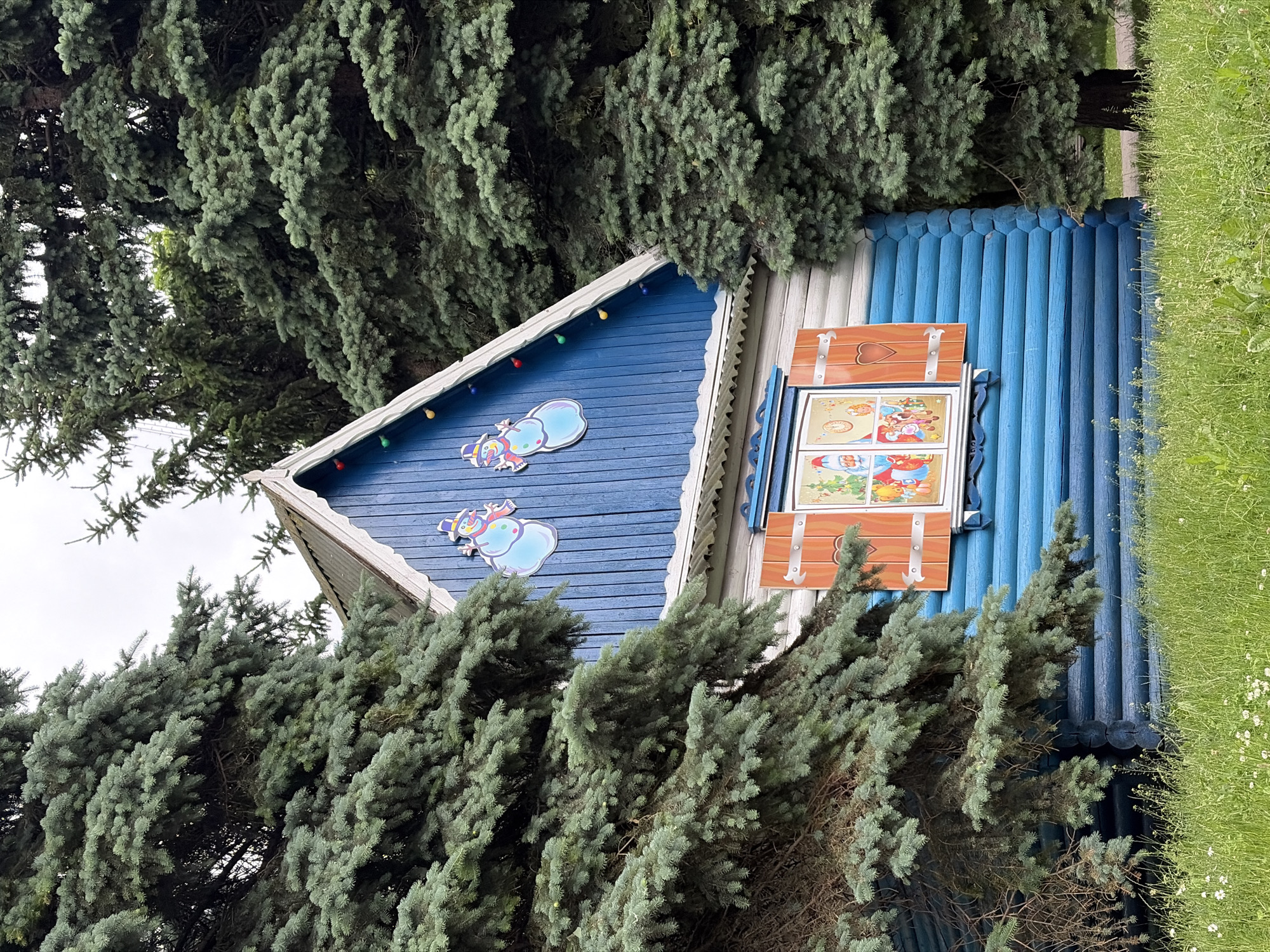 A wooden house with blue siding and a peaked roof decorated with festive lights is partially obscured by large evergreen trees. Two cartoonish snowmen are affixed to the upper blue portion of the house. A window with orange shutters is visible on the lower blue section of the house, and the glass of the window contains a colorful scene with figures that appear to be holiday-themed characters. The house is situated in a grassy area with small white flowers, and a paved path is visible to the right of the house. This structure is located at the "Santa Claus's Residence" in the Loshitsa Park, Minsk, Belarus.