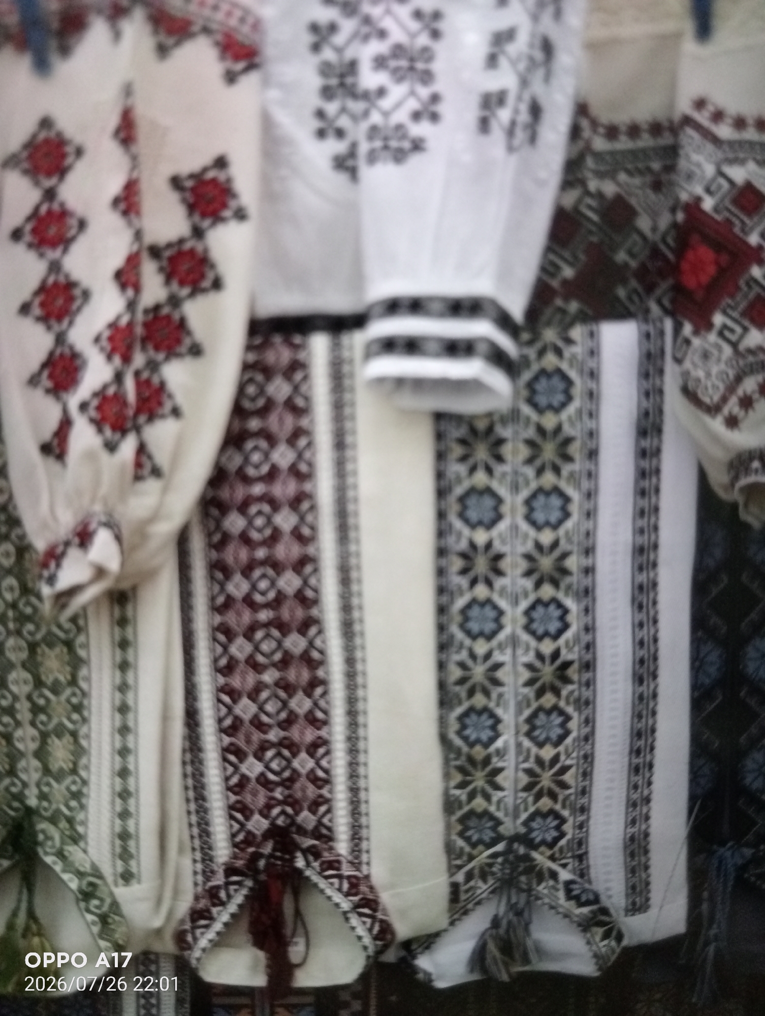The image shows a close-up, vertically oriented view of several embroidered garments, likely traditional folk clothing, displayed in what appears to be an outdoor market or stall. The embroidery features intricate geometric patterns and floral motifs in colors such as red, black, white, green, and maroon. The garments are made of a light-colored fabric, possibly linen or cotton. The overall impression is one of traditional craftsmanship and cultural heritage. The timestamp on the image indicates it was taken on July 26, 2026, at 22:01, and includes the text "OPPO A17," suggesting it was captured with a mobile phone. The scene is located in the city of Құмөзек, Kazakhstan.