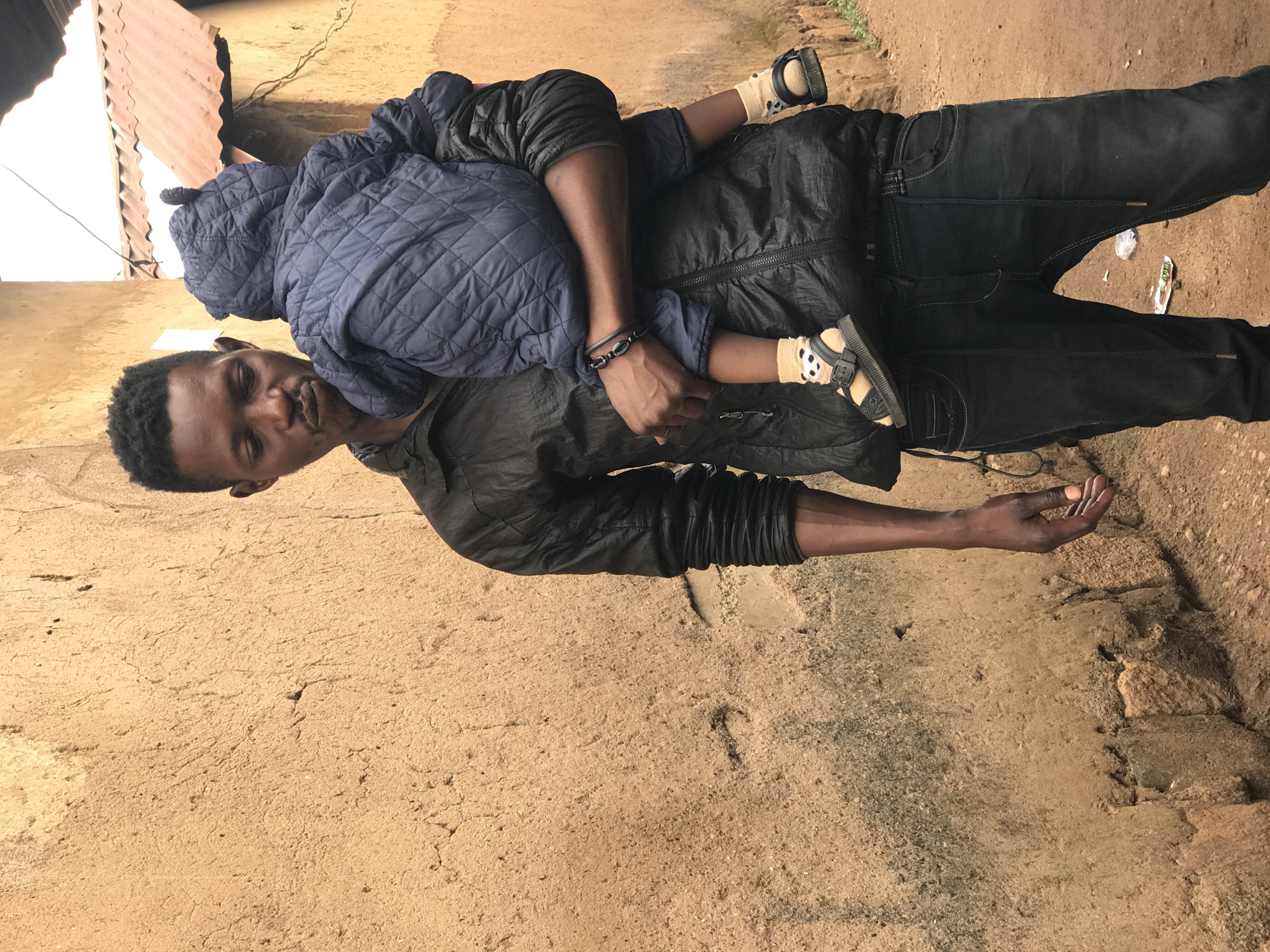 In Kabong, Nigeria, a man stands outdoors, carrying a young child. The man, with short, dark hair and a focused expression, wears a black, quilted jacket and dark jeans. The child, wrapped in a quilted blue jacket with a hood, is held in the man's arms. The child's feet are visible, wearing socks with panda designs and brown sandals. Behind them, a textured, beige wall dominates the background, showing signs of age and repair. Above the wall, a corrugated metal roof with a reddish-brown hue is partially visible. The ground beneath them is a dusty, reddish-brown earth.