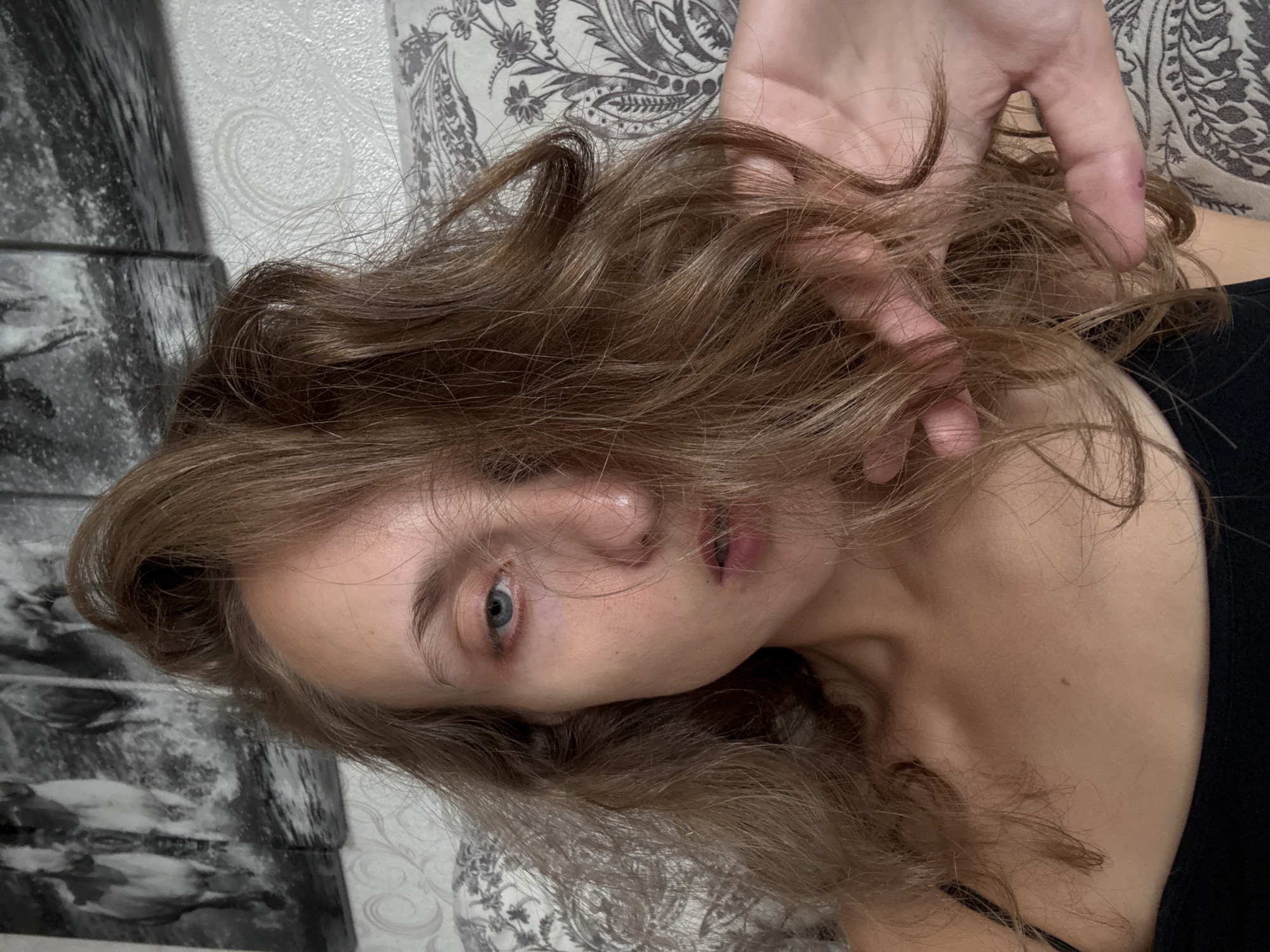 A close-up selfie shows a young woman with light brown hair, her face partially obscured by her hair. She is wearing a black tank top. The background consists of a wall with textured wallpaper and a framed artwork depicting horses. The image appears to be taken indoors, possibly in a bedroom.