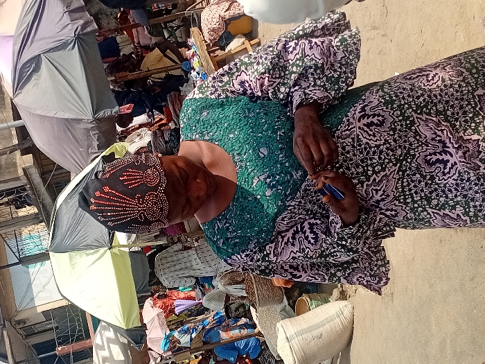 The image depicts an outdoor market scene in Ogaminan, Nigeria. In the foreground, an adult female with dark skin is central, looking downwards. She wears a long-sleeved dress featuring a green, purple, and white patterned fabric, and a black headwrap adorned with symmetrical golden-orange beadwork in a fan-like design. Her hands are positioned near her chest; she holds a small blue object in her left hand and is actively manipulating a small, thin object with her right hand.
The background reveals a bustling market environment with numerous individuals, most partially obscured. Multiple large umbrellas provide shade, including a prominent black and light green umbrella directly behind the primary subject. Market stalls with various goods are visible, and a large white sack, likely containing bulk items, rests on the ground to the left. Woven baskets are discernible among the merchandise. Buildings are visible in the distant background, and the ground appears to be a paved surface.