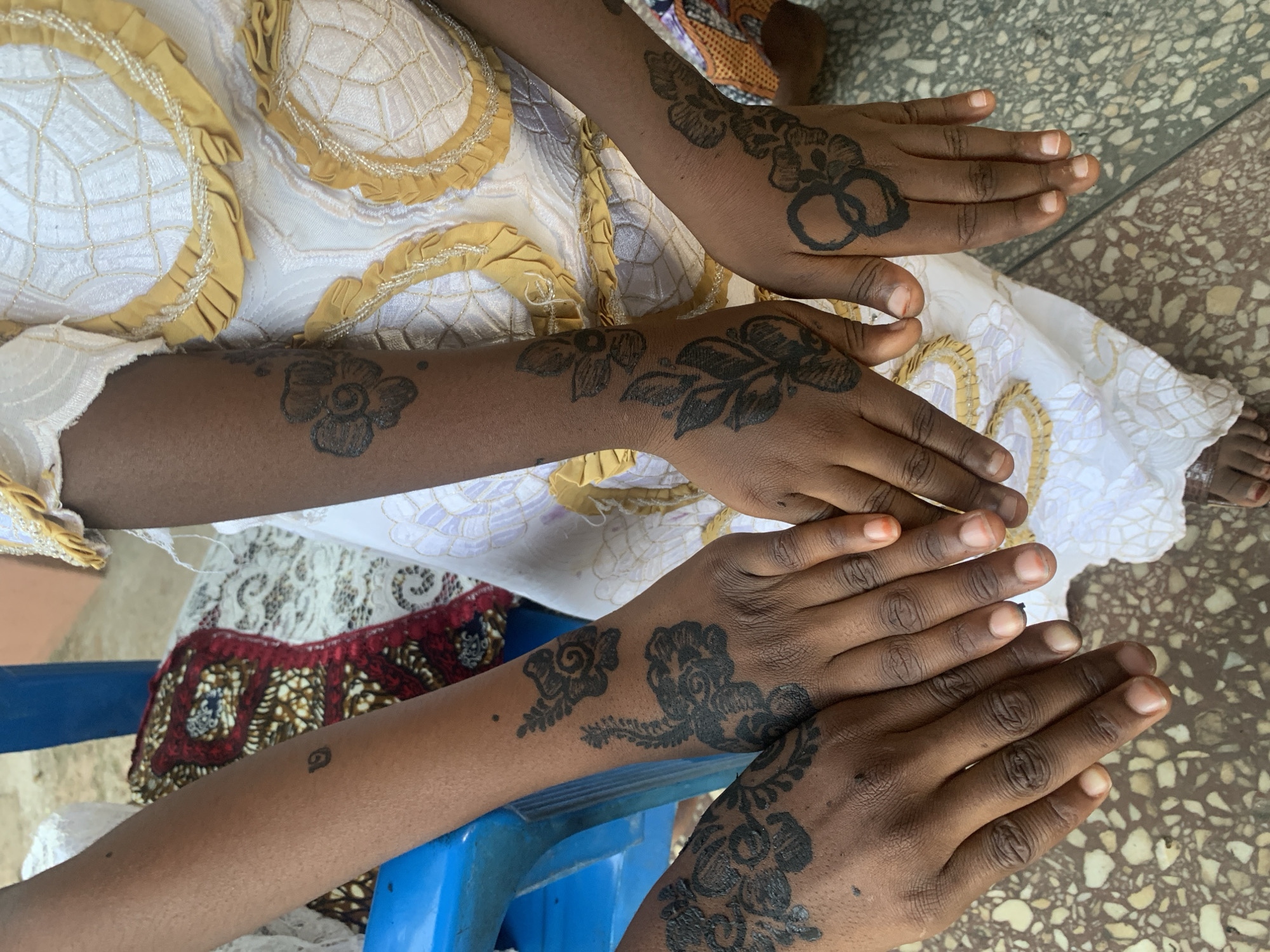 Three dark-skinned individuals are seen in the image, showcasing henna tattoos on their hands and arms. The tattoos feature floral and geometric designs. One individual is wearing a white and yellow dress with ruffled detailing. A blue plastic chair is partially visible. The floor appears to be tiled in a speckled pattern. The location is likely Jos, Nigeria, given the style of dress and henna application, which are common in the region.