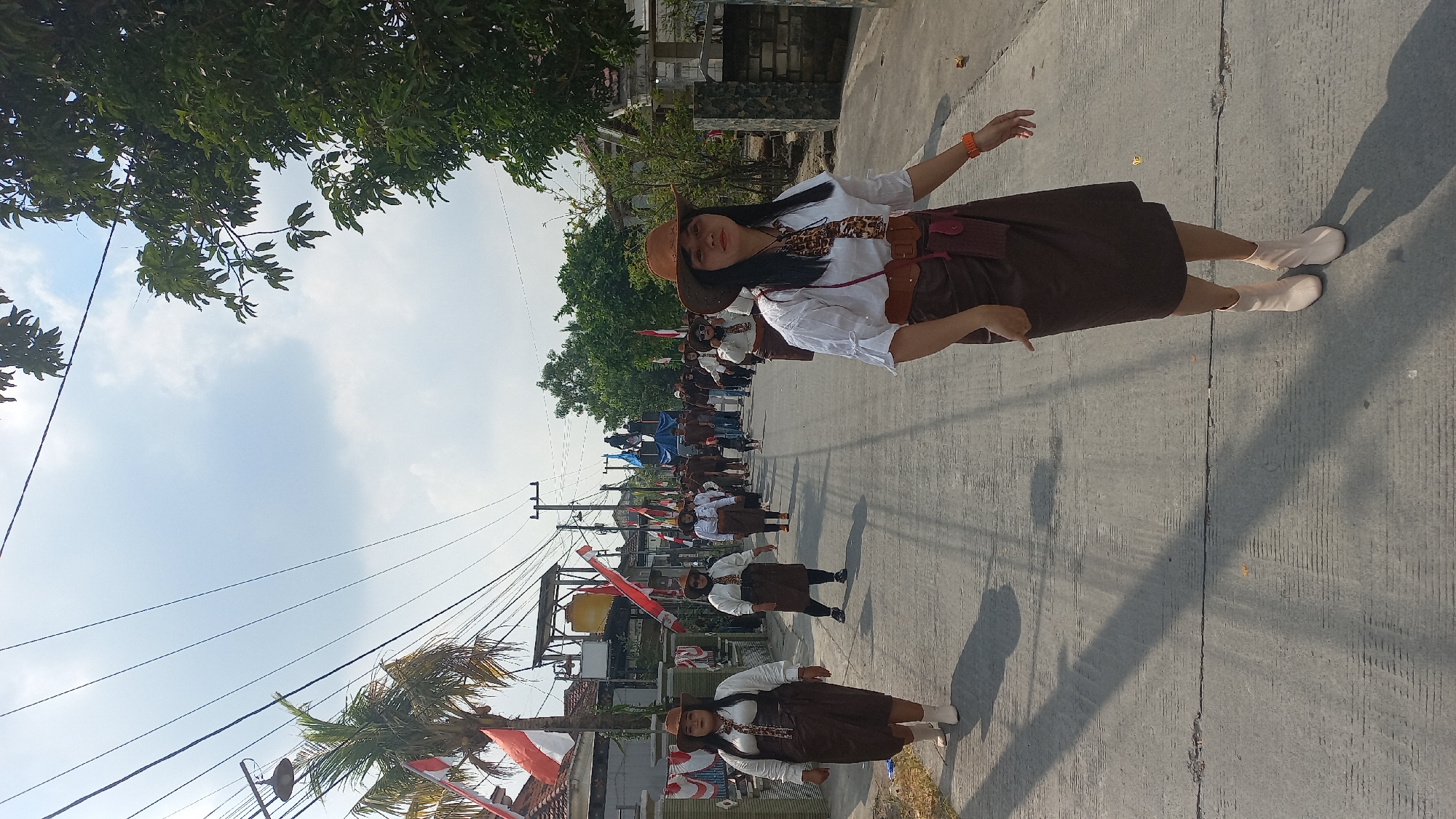 In Kedungmegarih, Indonesia, a group of women in matching brown skirts, white shirts, and cowboy hats participate in a parade along a paved street. The women are arranged in staggered lines, walking towards the viewer. The woman in the foreground on the right wears a brown belt and a tan lanyard around her neck. The street is lined with houses and trees. Indonesian flags are visible on poles along the sides of the street, and overhead wires stretch across the sky, which is partly cloudy. In the distance, a truck with people on top is part of the parade.
