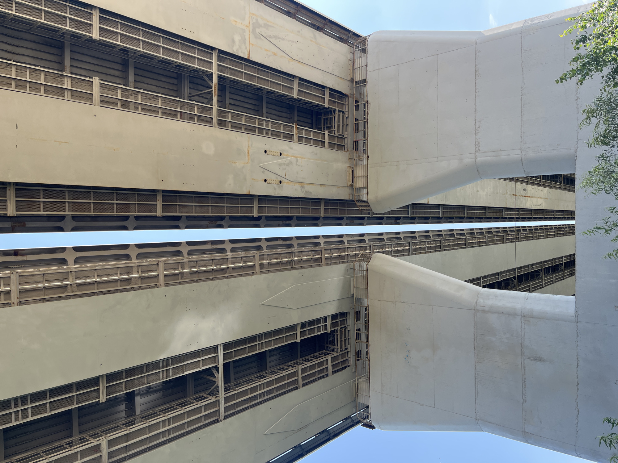 An underside view of a concrete bridge in Ufa, Russia, with a clear blue sky visible above. Two massive concrete pillars support the bridge deck. The bridge deck itself is made of large concrete slabs with an intricate metal support structure visible underneath. The perspective is looking upwards from below the bridge. In the lower right corner of the image, some green foliage from trees is visible.