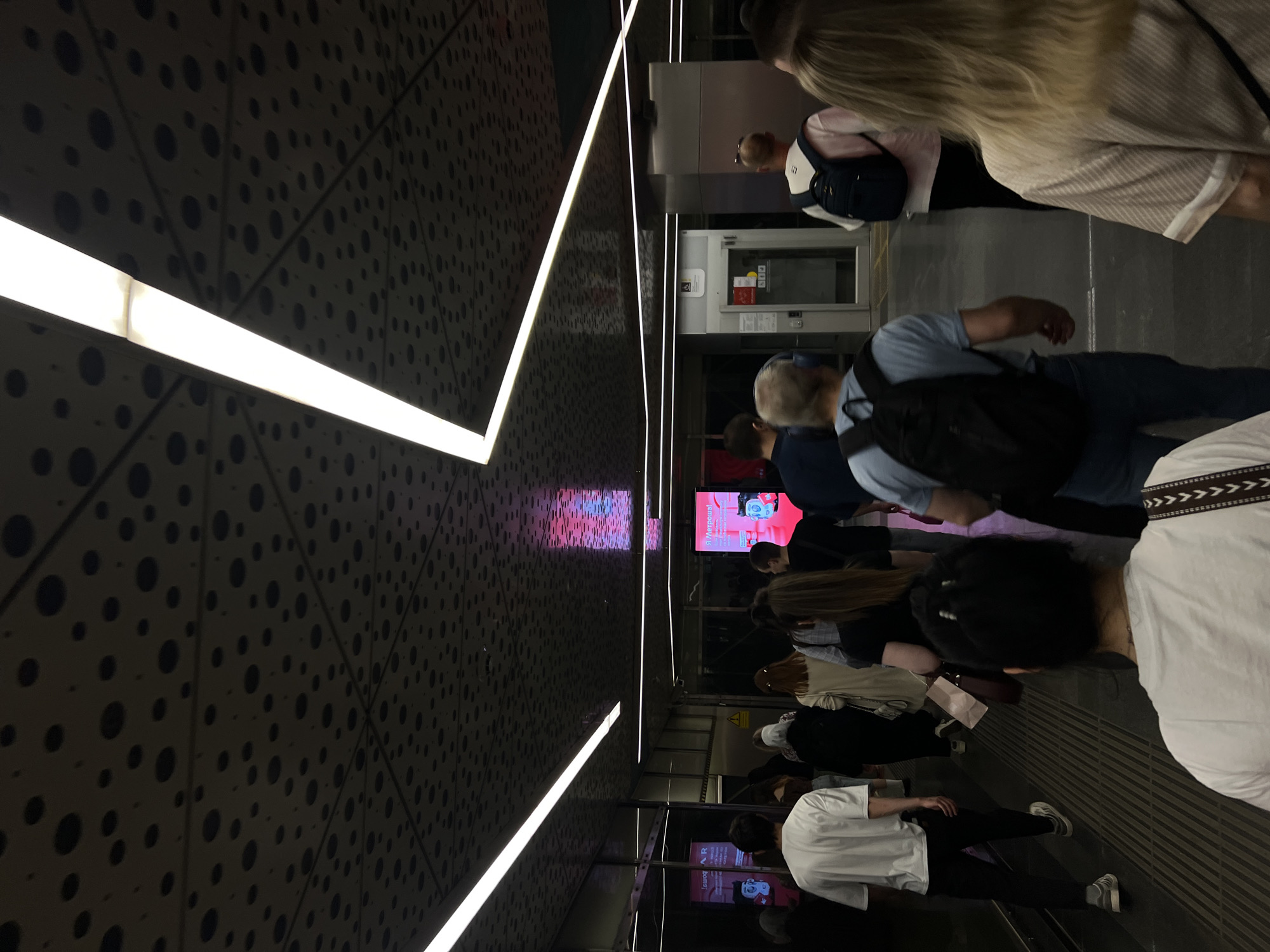 The image shows a dimly lit interior, possibly a subway station in Moscow, Russia, suggested by signage in the background. The ceiling and walls feature a geometric design with dark circles against a lighter gray background, emphasized by linear fluorescent lights. A group of people, mostly adults, are walking in the same direction. Their attire consists of casual clothing. Some are wearing backpacks. Most are partially obscured or viewed from the back. In the background, screens are visible, displaying advertisements or public information. The floor is gray.