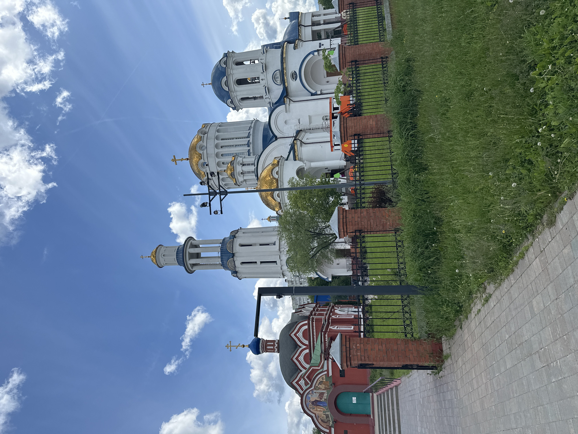 The image shows the Novodevichy Convent in Moscow, Russia, from a ground-level perspective, rotated 90 degrees counterclockwise. The architecture features multiple churches with white walls, blue and gold domes adorned with Orthodox crosses. A decorative red brick building with a green door is also visible. A black metal fence encloses a grassy area in front of the buildings. Paved pathways and patches of blue sky with white clouds are present. The image was taken during daylight hours. No people are visible.