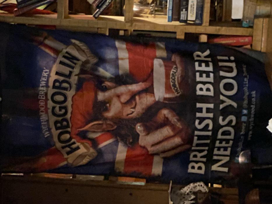 A banner hangs against a background of wooden shelves filled with books and other items. The banner features a goblin-like figure with pointed ears and a red cap, superimposed on a stylized Union Jack flag. The words "HOBGOBLIN" appear in large letters along with "WYCHWOOD BREWERY." To the right, the text "BRITISH BEER NEEDS YOU!" is prominently displayed. The banner also includes website and social media information.
