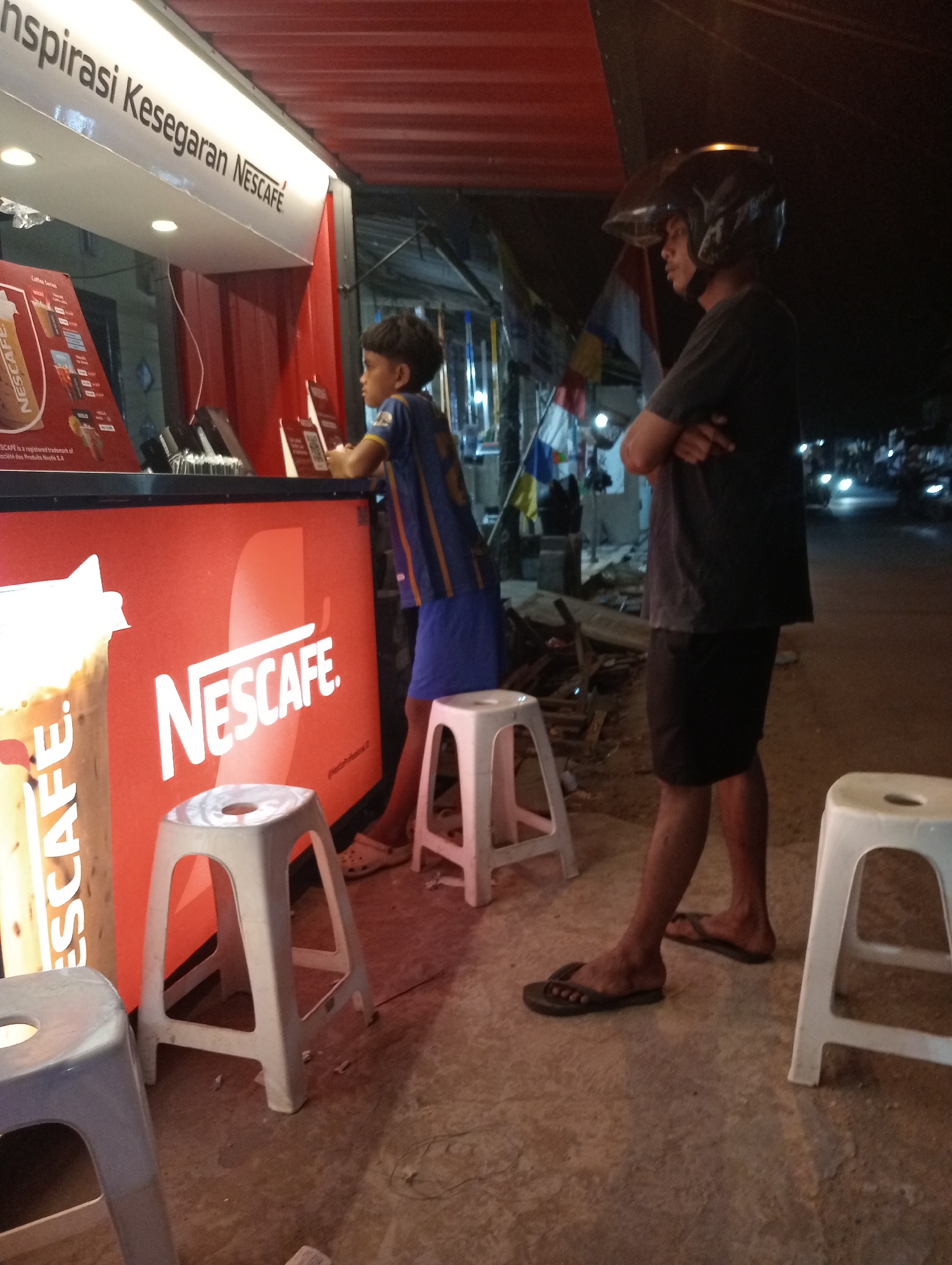 The image depicts a nighttime scene at a Nescafé beverage stand, likely located on a street in Jambi, Indonesia, given the context provided. Two individuals are present. A younger male, wearing a blue and yellow shirt and shorts, stands at the counter, looking towards the right. An adult male, wearing a dark t-shirt, dark shorts, and a motorcycle helmet with the visor up, stands to the right of the younger male with his arms crossed. He is looking towards the right as well. In the foreground, there are several white plastic stools. The Nescafé stand itself is red with the brand logo prominently displayed. The background suggests a street environment with blurry lights and other figures, indicating it is an active area at night.