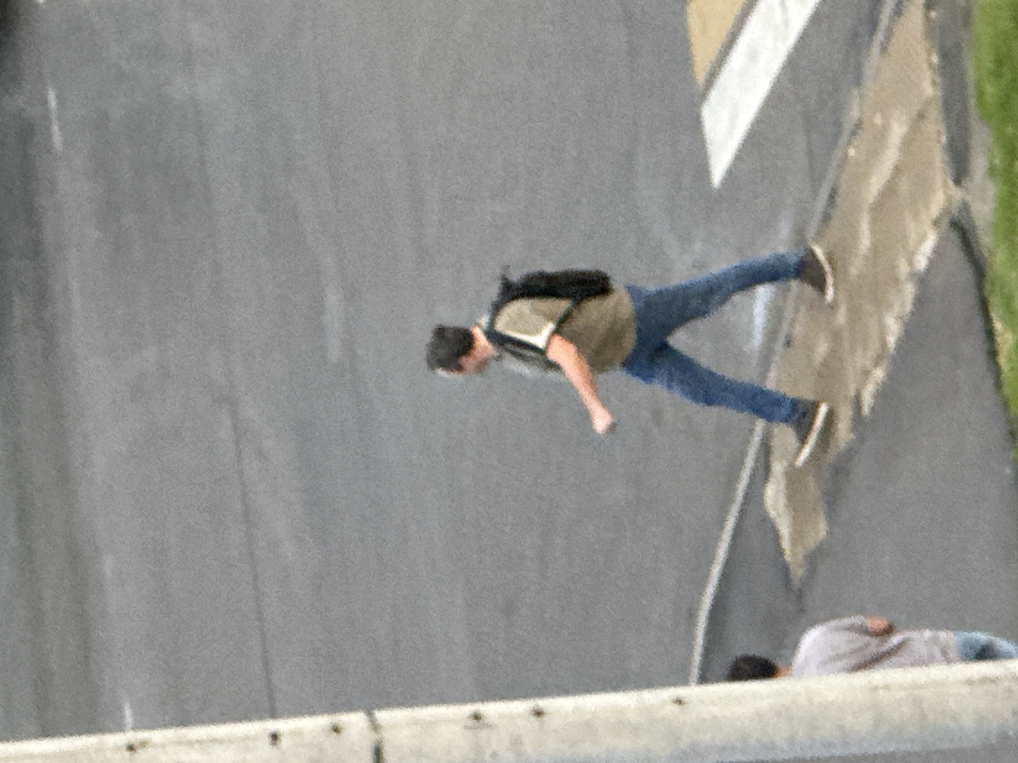 A man wearing a khaki shirt, blue jeans, and a black backpack walks across a crosswalk. He is on a paved street in Podolsk, Russia. Another person, only partially visible, stands to the left of the frame, wearing a grey t-shirt and blue jeans. A white pole, possibly a light post or signpost, is visible on the far left of the image. The ground is a mixture of asphalt and concrete.