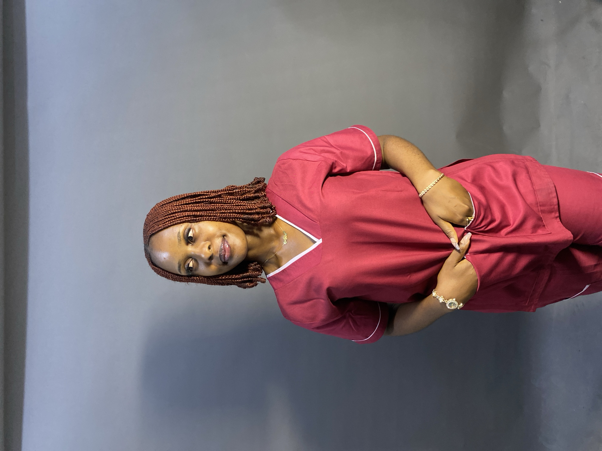 A woman with brown braided hair is posing in a red scrub suit with white trim against a dark gray wall. She is wearing a gold bracelet and necklace. The photograph appears to have been taken indoors with artificial lighting. There is no information that would confirm the location in the city of Jos, Nigeria.