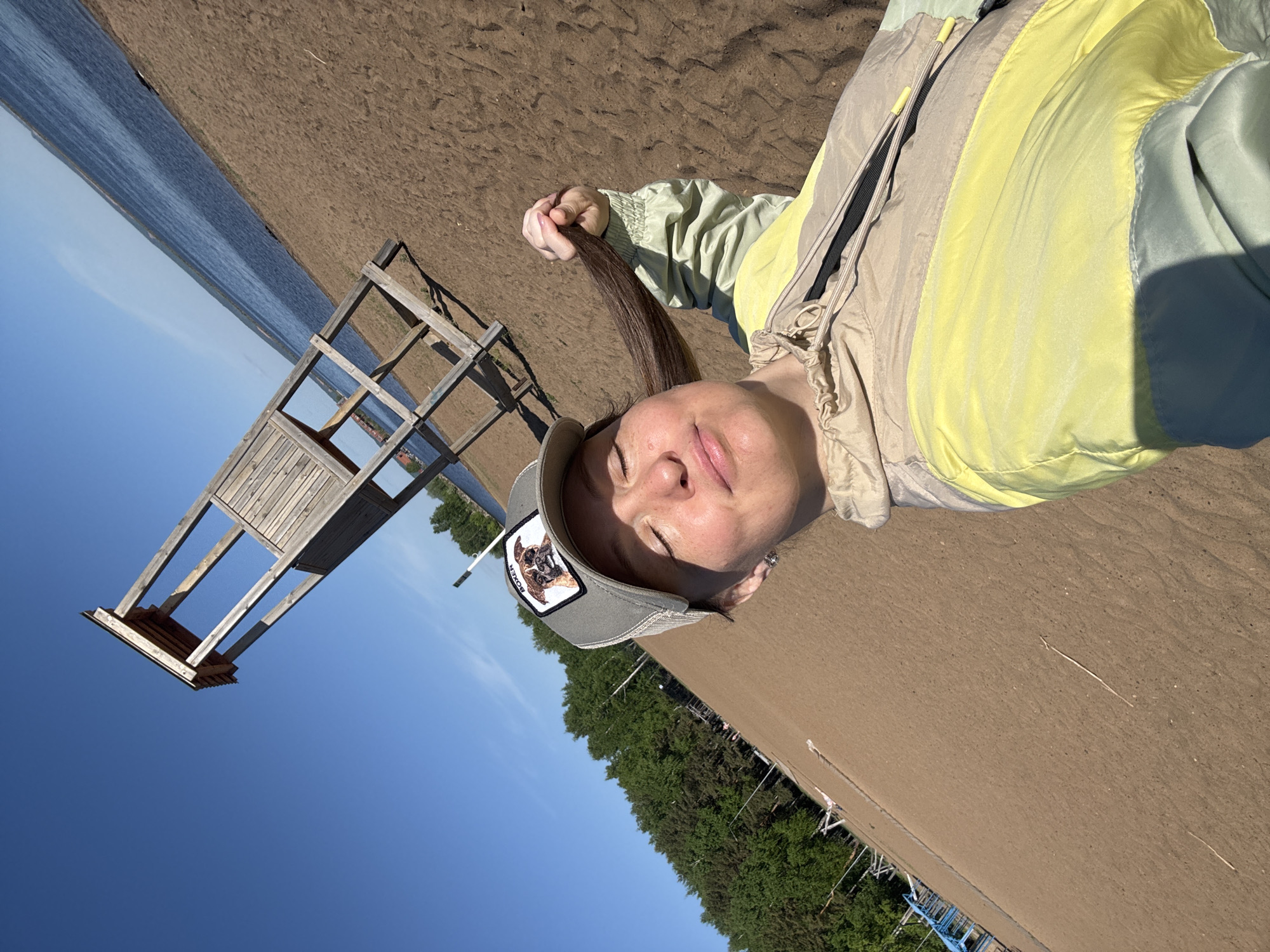 A woman wearing a baseball cap and a yellow jacket lies on a sandy beach in the city of Chaikovsky, Russia, located by the Kama River. She is holding a strand of her brown hair. A lifeguard tower stands on the sand near the water's edge, while a line of trees borders the other side of the beach. The sky is clear and blue.