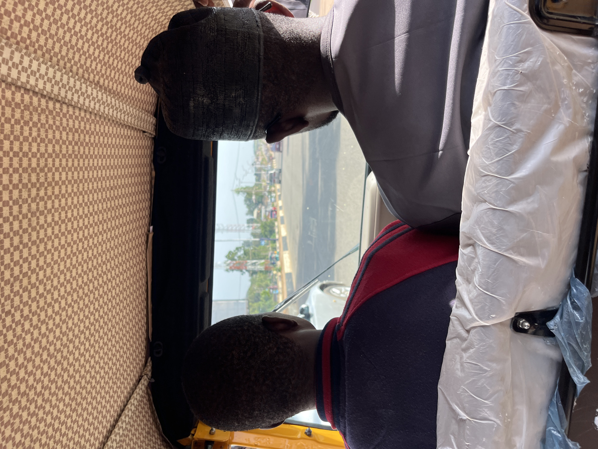 An overhead shot captures the interior of a tuk-tuk vehicle traveling on a paved road in Bukuru, Nigeria. Two individuals are seated in the front, facing forward. The person on the left, wearing a dark blue and red shirt, has their back to the camera. To their right, a person wearing a gray outfit and a dark cap is also facing forward. The tuk-tuk's ceiling is covered with a patterned beige fabric. Through the windshield, a street scene is visible with other vehicles, buildings, and utility poles. The seats in the foreground are covered with white plastic sheeting.