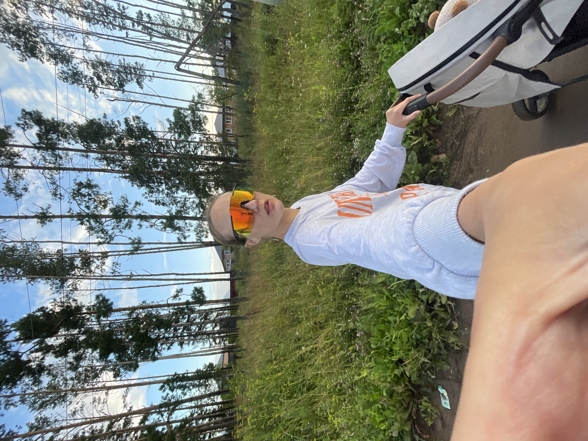 In an outdoor, medium shot, a young person wearing oversized orange mirrored sunglasses and a light gray long-sleeved sweatshirt with orange lettering is pushing a white baby stroller. The stroller has a brown handle and a teddy bear can be seen inside. The person is on a dirt path next to a field of tall green grass and wildflowers. Behind the field, a line of tall, slender pine trees borders the scene. In the distance, behind the trees, several brick houses with white trim are visible, suggesting a rural or suburban setting. The sky is blue with scattered white clouds. The lighting suggests it is daytime, likely late afternoon given the angle of the sun. The image appears to be a selfie, as the person's arm and hand are prominently in the foreground. The overall mood is casual and perhaps a little adventurous, due to the bold sunglasses. No visible text other than the lettering on the sweatshirt. The location is identified as Podshivalovo, Russia.