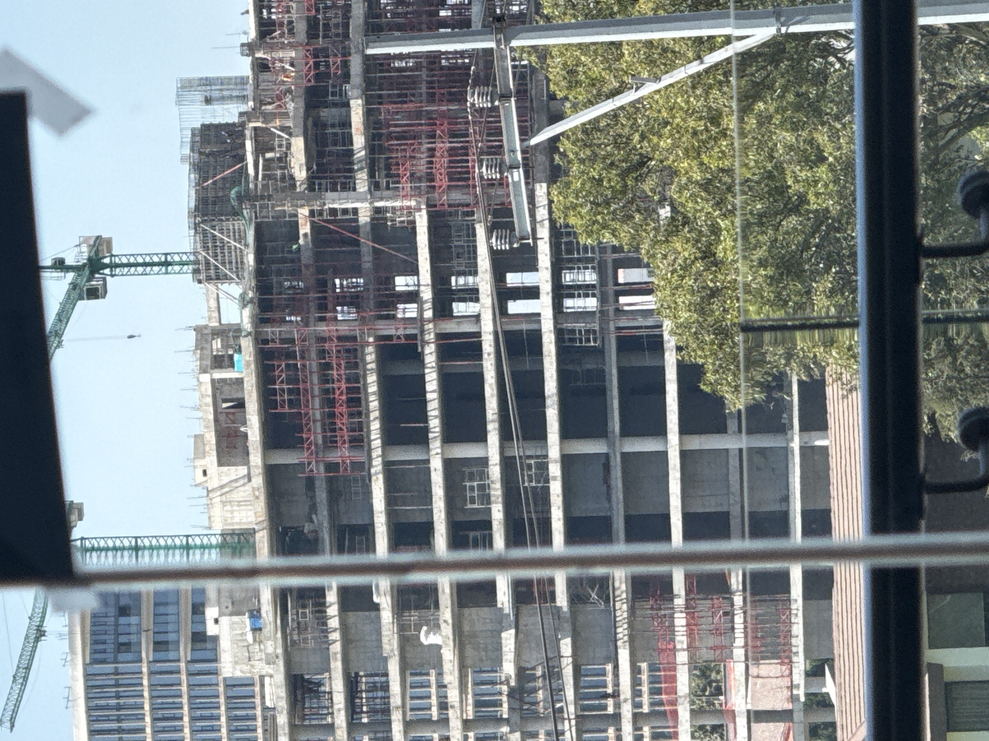 The image depicts a large, multi-story building under construction, viewed from an elevated perspective, likely through a window given the reflections and foreground elements. The scene is set in San Bernardino Tlaxcalancingo, Mexico, during daytime with a clear blue sky.
The primary subject is the skeleton of a modern high-rise building, primarily composed of reinforced concrete columns and floor slabs. Extensive red metal scaffolding covers various sections of the building's exterior and interior, indicating ongoing construction work. A large green tower crane is prominently visible on the left side of the frame, its jib extending horizontally across the site, suggesting heavy lifting operations are underway.
To the right of the construction site, a large, leafy tree with dense green foliage partially obscures the building, providing a natural contrast to the industrial scene.
The foreground shows elements of a window frame or railing, which are out of focus and create reflections, particularly noticeable as a horizontal line and vertical bar across the lower and right portions of the image. These reflections, along with the out-of-focus elements, suggest the photograph was taken from indoors, looking out.
No people are clearly visible, and there is no discernible text within the image. The overall impression is one of active urban development under fair weather conditions.