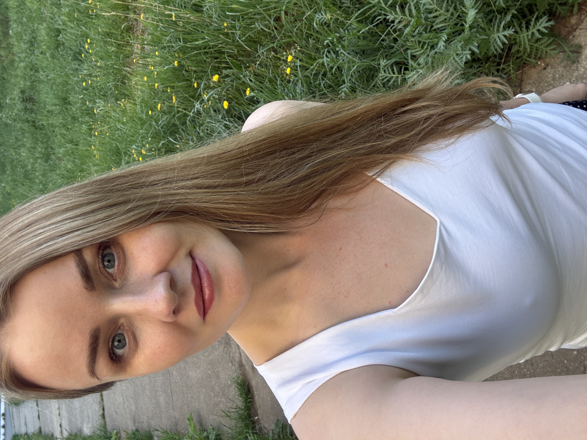 A woman with blonde hair and light complexion stands outside. She is wearing a white tank top. Behind her, there is green grass and yellow flowers. There is a concrete surface in the background. The location is likely in the city of Chaikovsky, Russia.