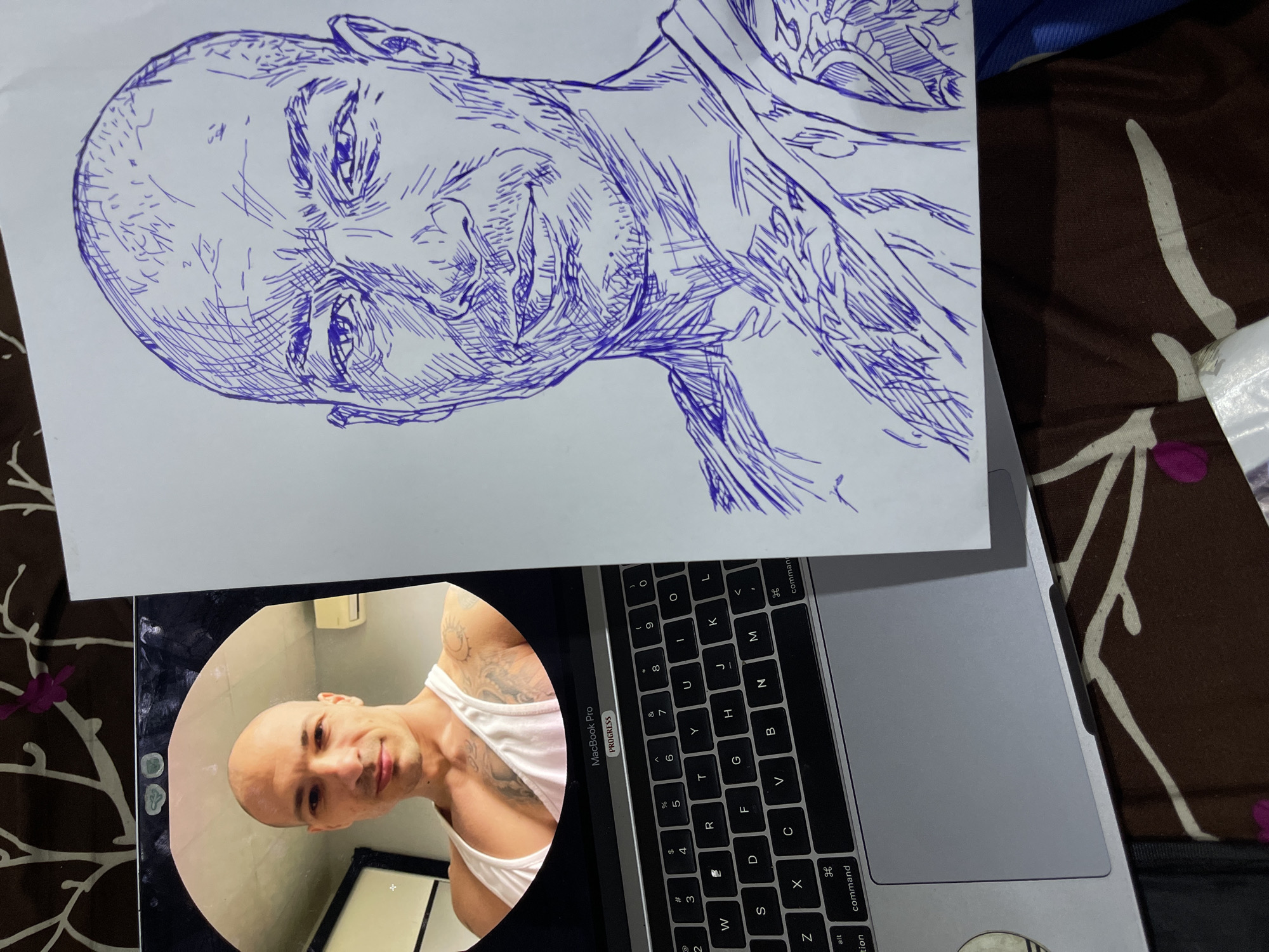 A pen-and-ink sketch of a bald man's head and shoulders is displayed on a white sheet of paper, resting on a brown and white patterned fabric. To the left of the sketch, a MacBook Pro laptop is partially visible. A circular photo of the same bald man, wearing a white tank top and showing tattoos on his chest and arm, is displayed on the laptop screen. The photo shows the man from the chest up, with a white wall and an air conditioning unit in the background. The scene is illuminated by overhead lighting. The sketch captures the man's facial features and the outline of his clothing, with blue ink used for all lines and shading. The laptop's keyboard is visible, featuring standard QWERTY layout.