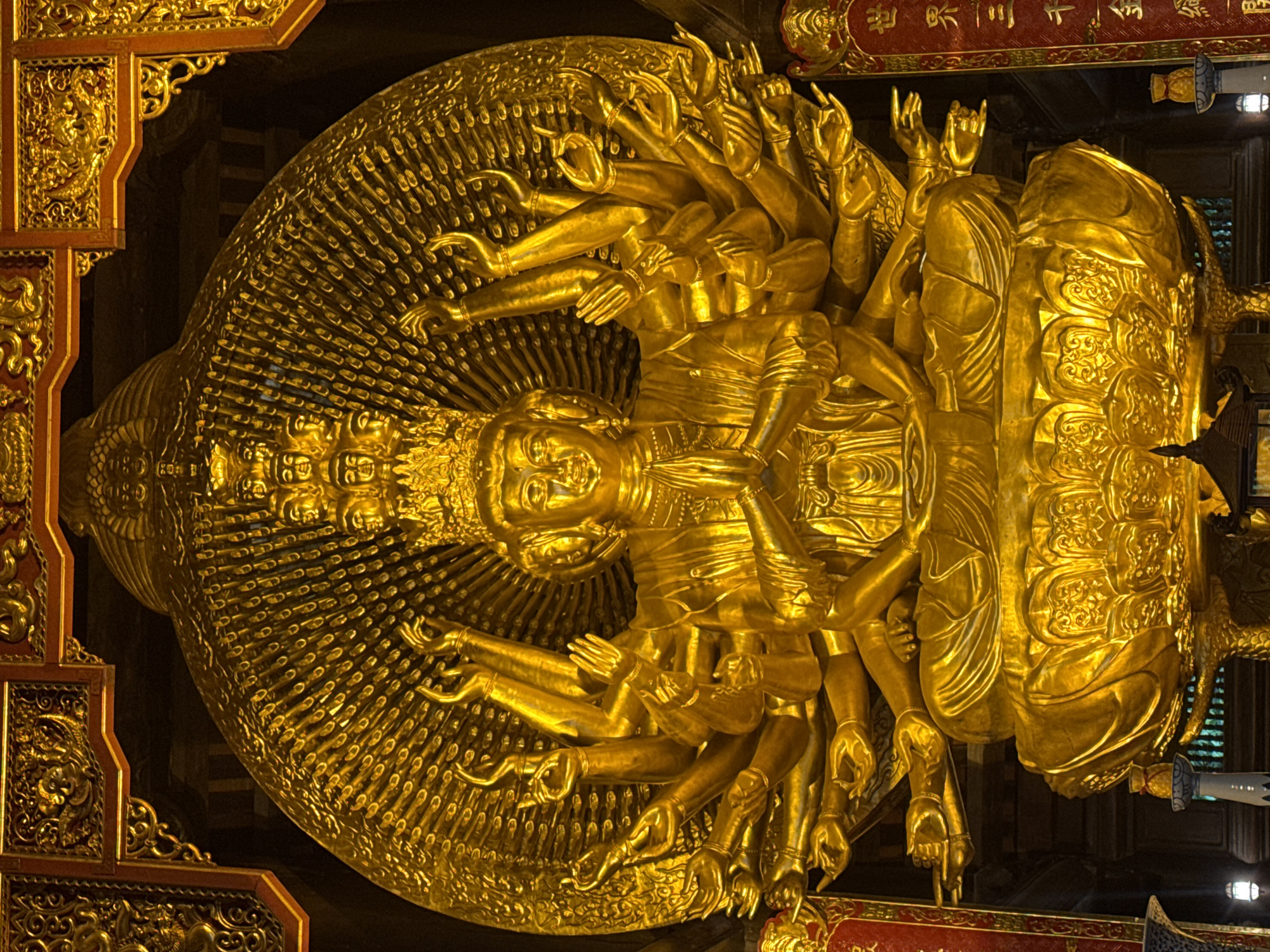 The image shows a large, ornate golden statue of a multi-armed deity, likely a Buddhist figure like Guanyin, with numerous hands. The statue is seated on an elaborate lotus base, with a radiating halo behind it, also adorned with many smaller faces. The deity wears a crown and is depicted with hands in various mudras (symbolic hand gestures). The statue is bathed in golden light, highlighting its intricate details. In the background, elements of a temple or shrine are visible, including carved wooden structures and red banners with gold lettering. This statue is located within the Bích Động Pagoda complex in Ninh Bình province, Vietnam, which is near the ancient capital of Tây Hoa Lư Ward.