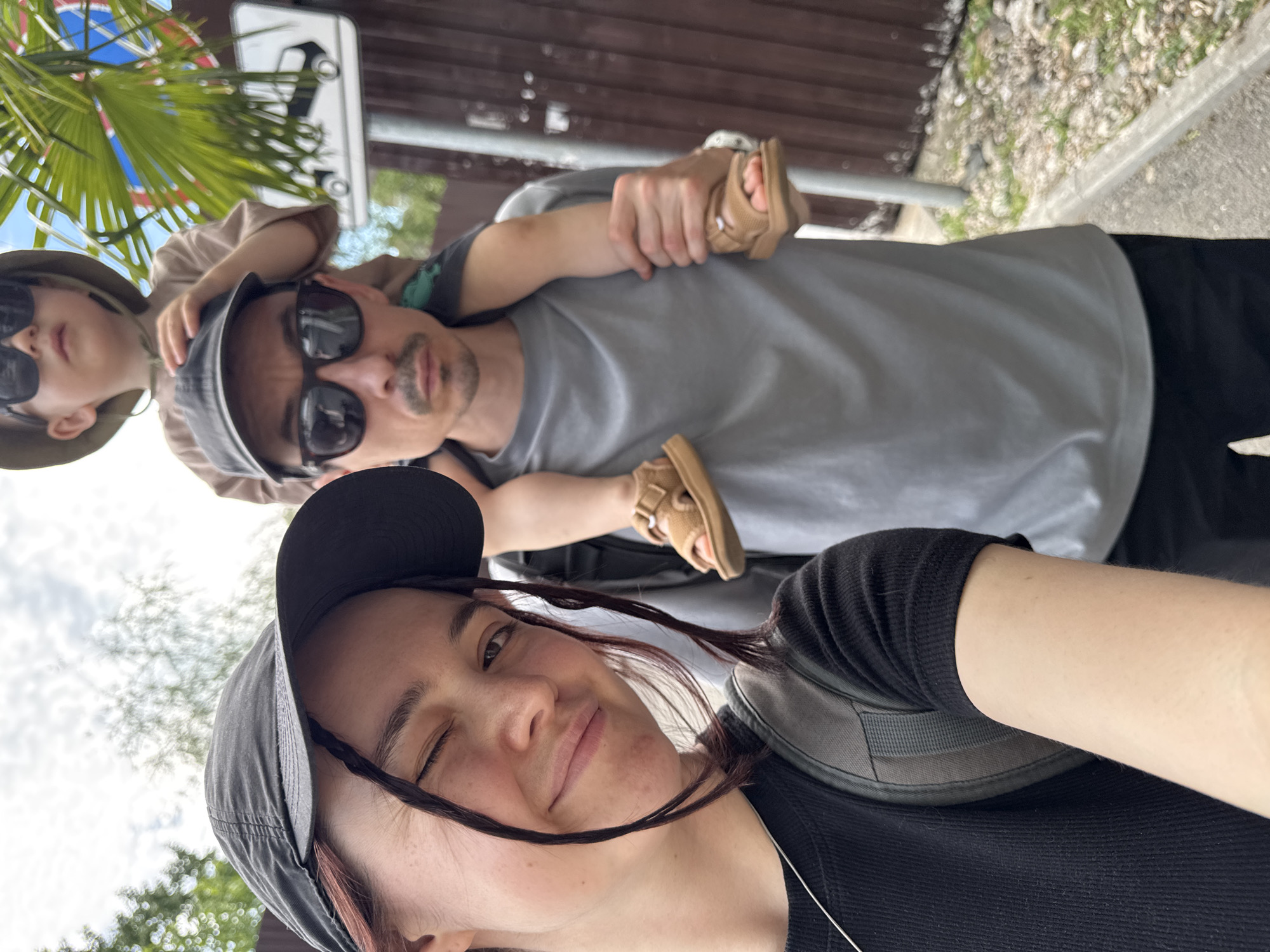 A selfie shows a woman in the foreground wearing a black baseball cap and a black shirt, winking with one eye open. Behind her, a man with sunglasses carries a child on his shoulders, also wearing sunglasses and a hat. The man is wearing a gray t-shirt. The child's sandals are visible behind the man's neck. The background includes greenery, a street sign, a wooden fence, and what appears to be a concrete path. The location is likely in Sirius, Russia.
