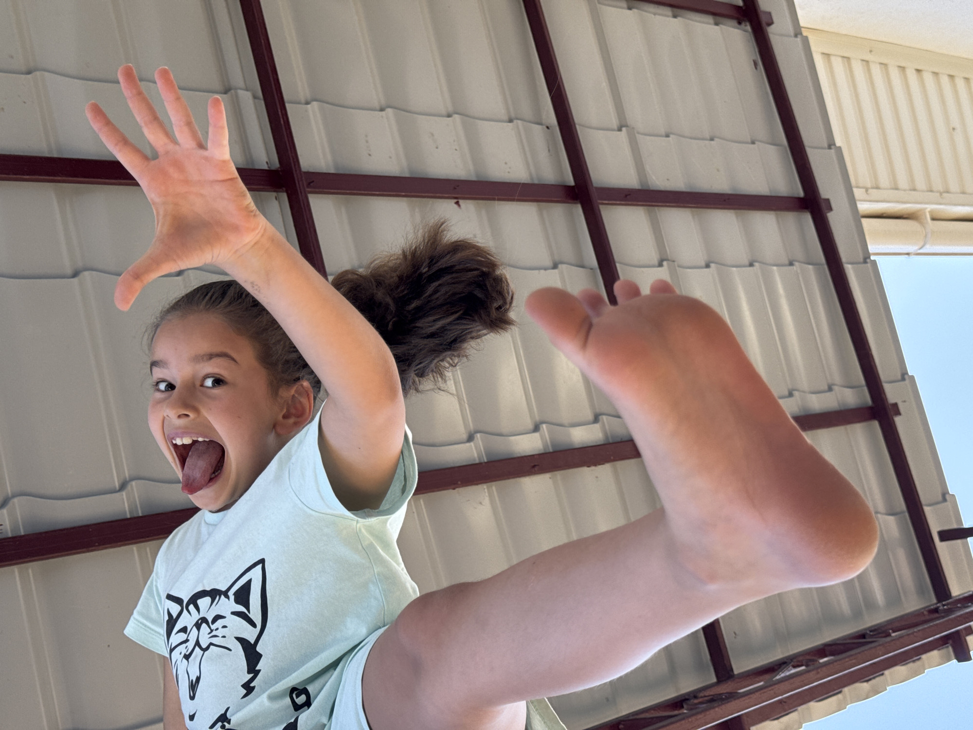 The media file features a young girl from a low-angle perspective, seemingly in motion. She is wearing a light green t-shirt with a black graphic of a cat with its tongue sticking out. Her left arm is raised with her hand spread wide, fingers extended, in a playful gesture. Her mouth is open wide, her tongue sticking out, suggesting excitement or a silly expression. Her right leg is extended upwards, with her bare foot prominently visible.
The background consists of a corrugated metal roof structure with dark brown beams. The sky is visible in the upper right portion of the image, indicating it is daytime. The lighting suggests it is a sunny day. The scene appears to be outdoors, possibly a backyard or recreational area, given the casual attire and the roof structure which could be part of a covered patio or shed. The overall mood is energetic and joyful, with the girl's expression and pose conveying a sense of fun and playfulness. The location context provided is Uyutnoye, Ukraine.