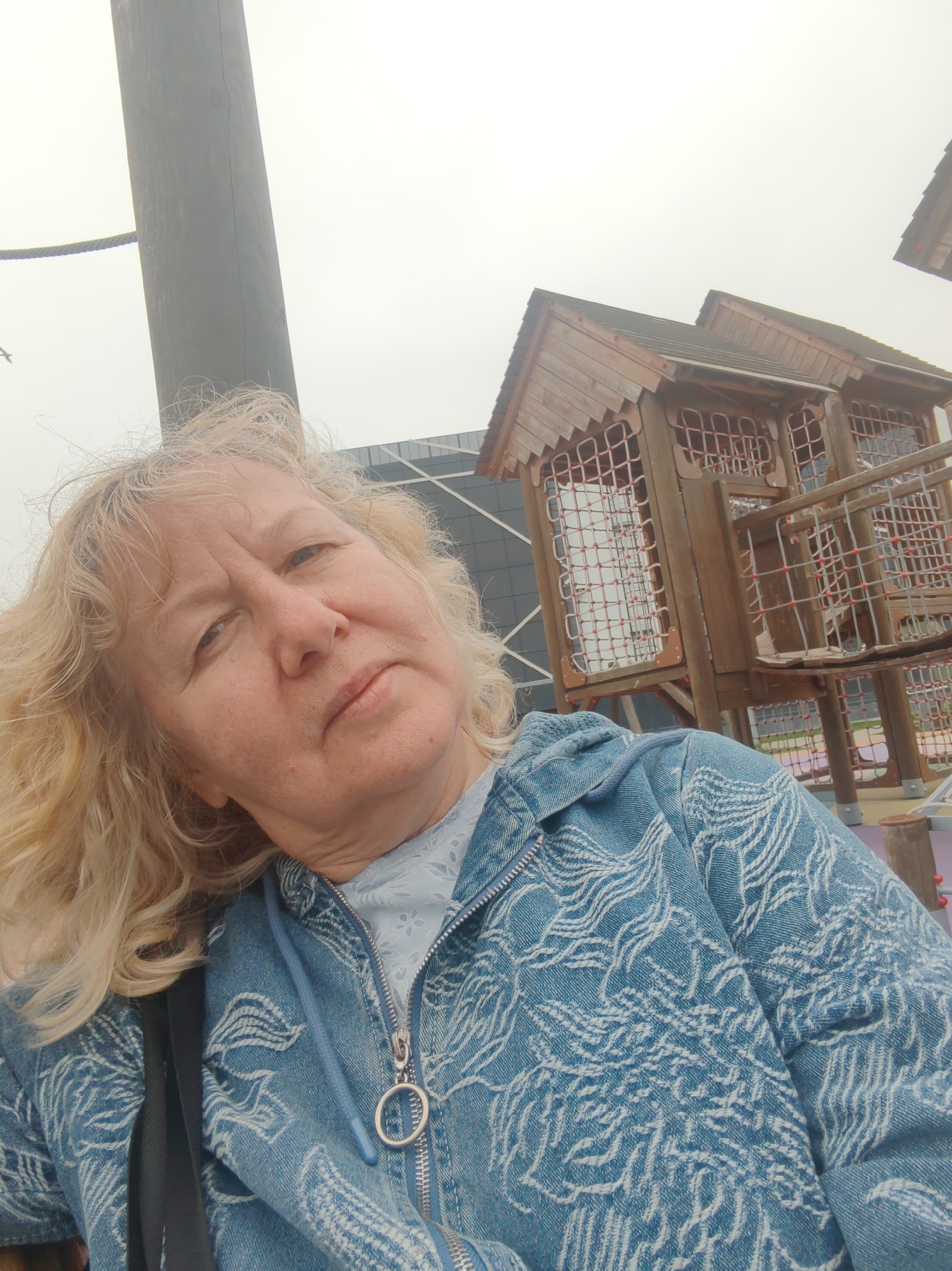 A woman with blonde, wavy hair is the subject of this medium-shot selfie, set against the backdrop of a children's playground. She is wearing a light blue zip-up jacket with a white patterned design.
Behind her, a complex wooden structure designed for children to climb and play on is visible. The structure is multi-tiered and features netted areas and platforms.
In the background, there is a modern building with glass panels, indicating the location is urban. The sky is overcast, lending a flat, muted light to the scene.
Based on the location, this photograph was taken at a playground in the city of Tver, Russia.