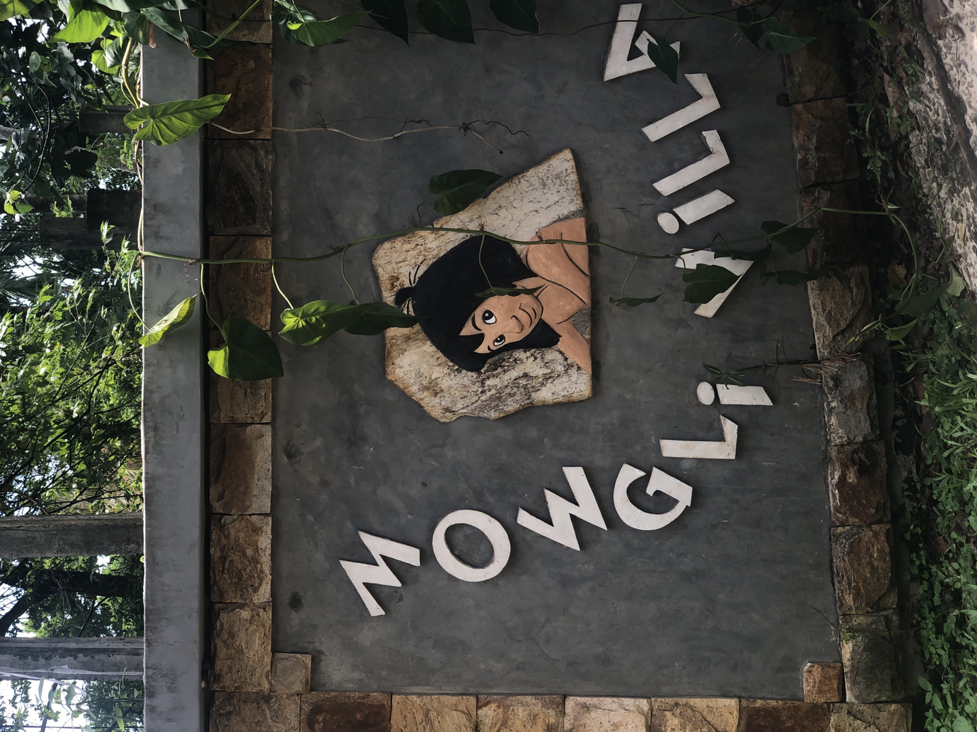 A sign reading "MOWGLI VILLA" is mounted on a grey concrete wall. The letters are white and appear to be made of wood or a similar material. To the right of the "MOWGLI" text, a cartoon depiction of Mowgli from The Jungle Book is painted on a rough, stone-like surface. The sign is partially obscured by green vines and leaves from surrounding foliage. The wall itself has a base of rough-hewn stones. The background shows trees and a bright sky, suggesting an outdoor, tropical location. The image captures the entrance to a villa or property.