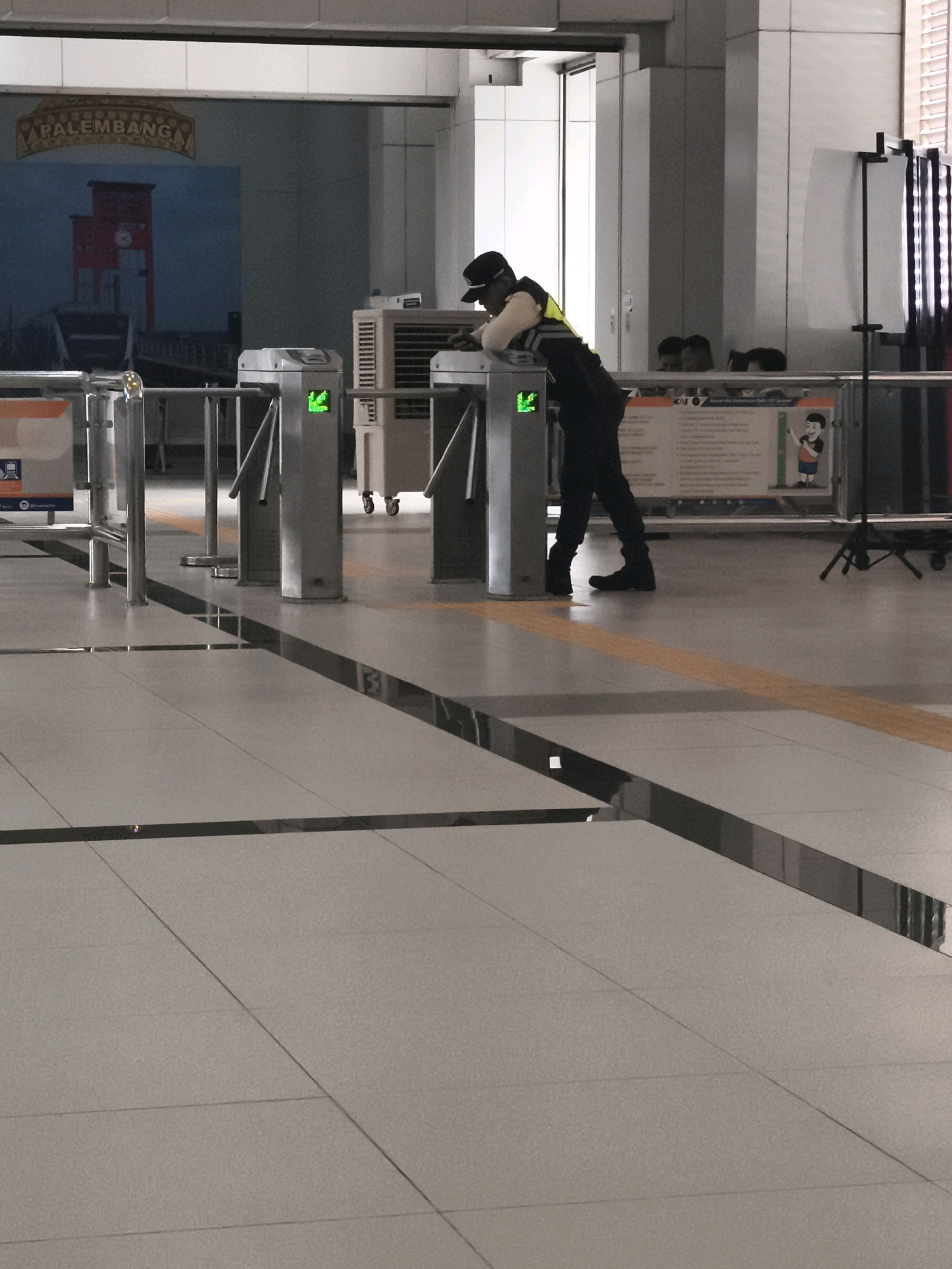 An image shows a security guard leaning on a turnstile in an indoor public space, possibly a train station, in Palembang, Indonesia. The guard is wearing a dark uniform with a yellow reflective vest and a cap. The turnstile has a green arrow indicating it is operational. In the background, a mural depicting a scene with a red building and boats is visible, with the word "PALEMBANG" prominently displayed. Other elements in the scene include an air conditioning unit, metal railings, and tiled flooring. There are also other people in the background, partially obscured, and informational posters. The overall lighting suggests an indoor environment with artificial illumination.