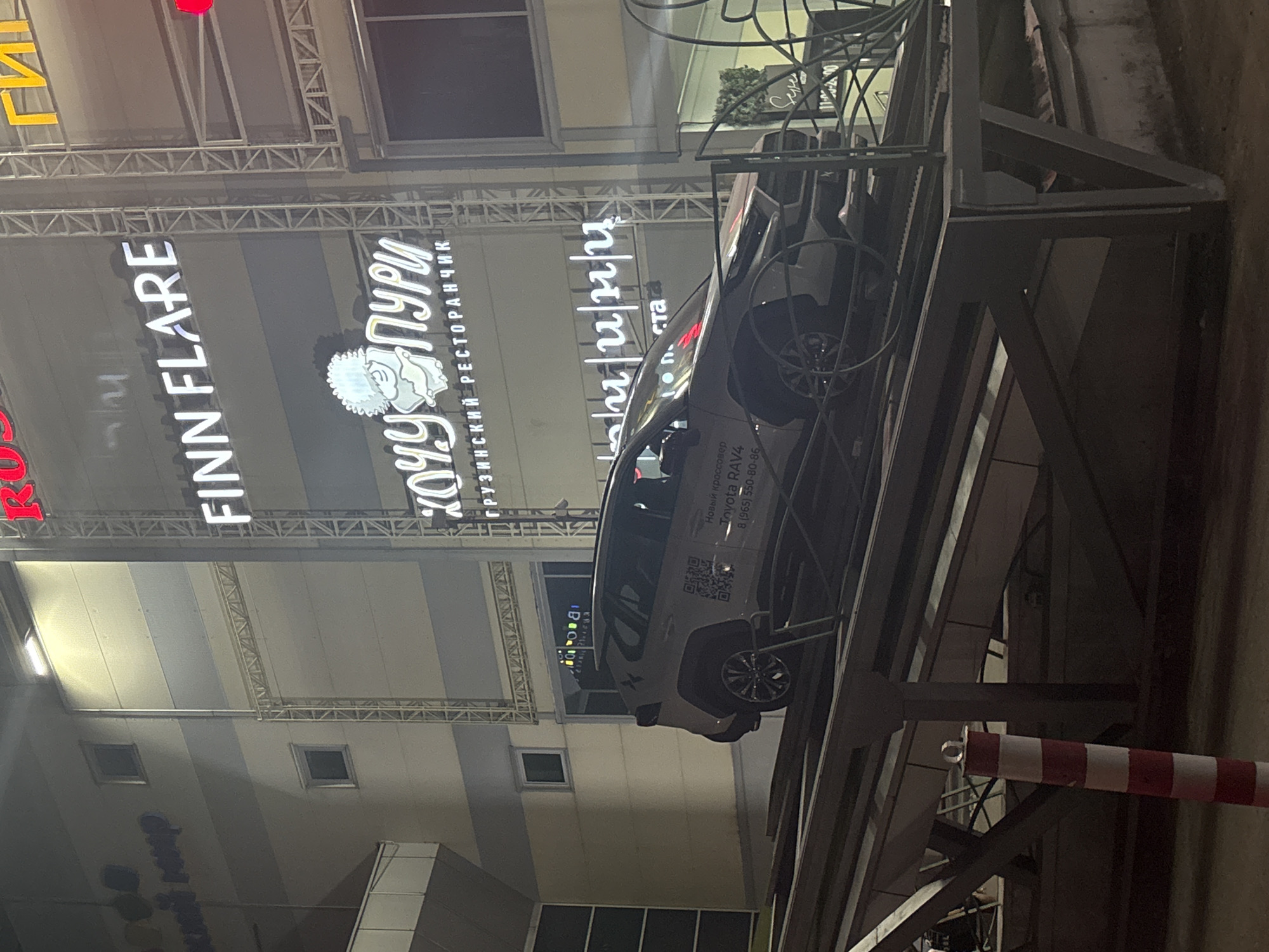 The image shows a white Toyota RAV4 vehicle, positioned on a metal structure, in what appears to be an advertising display. The vehicle has markings and QR codes on its side, along with the text "Новый кроссовер Toyota RAV4 8 (965) 550-80-86". The vehicle is angled and viewed from a low perspective.
In the background, large illuminated signs are visible. The sign on the left reads "FINN FLARE" in white letters. To the right of that is another sign in Russian, "ХОЧУ ПУРИ грузинский РЕСТОРАНЧИК", indicating a Georgian restaurant. The photo also depicts architectural elements of the building, with a grid-like metal framework in the background.
The scene is located in Perm, Russia.