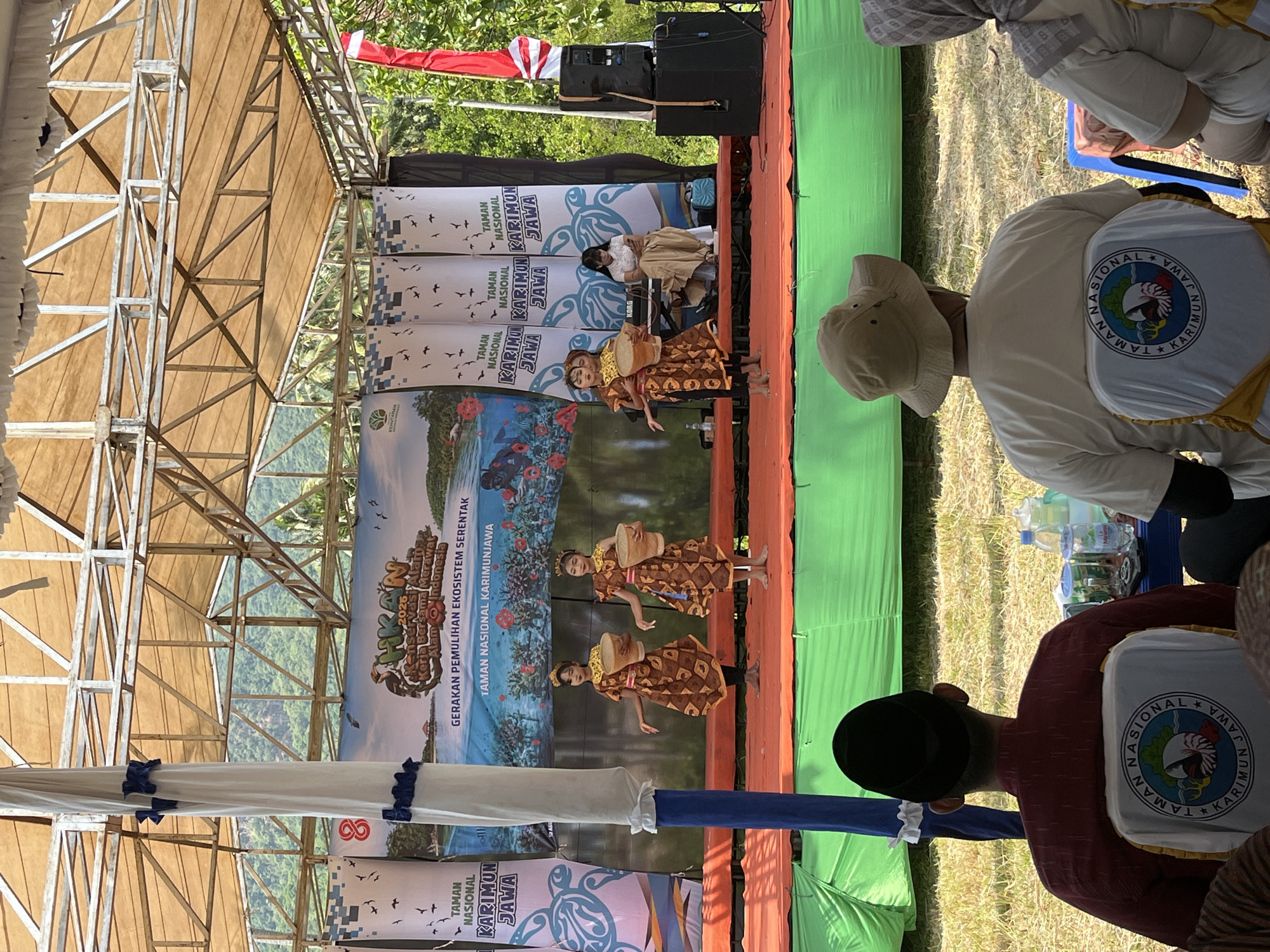 The image shows an outdoor stage event in Kemloko, Indonesia. On stage, two young girls in traditional brown and gold patterned dresses are performing a dance, each holding a woven basket. Behind them, a banner displays text related to "Gerakan Pemulihan Ekosistem Serentak Taman Nasional Karimunjawa." A woman in a white top and tan skirt is seated at a keyboard, possibly providing musical accompaniment. In the foreground, several people are seated in chairs, facing the stage. The chairs are adorned with white covers featuring a circular emblem with text that appears to say "Taman Nasional Karimunjawa." The environment is sunny with lush green foliage visible in the background, suggesting a tropical or subtropical location. The stage is set up under a large tent structure, and there are speaker systems on the right side of the stage. The overall scene depicts a community or official event focused on environmental conservation.