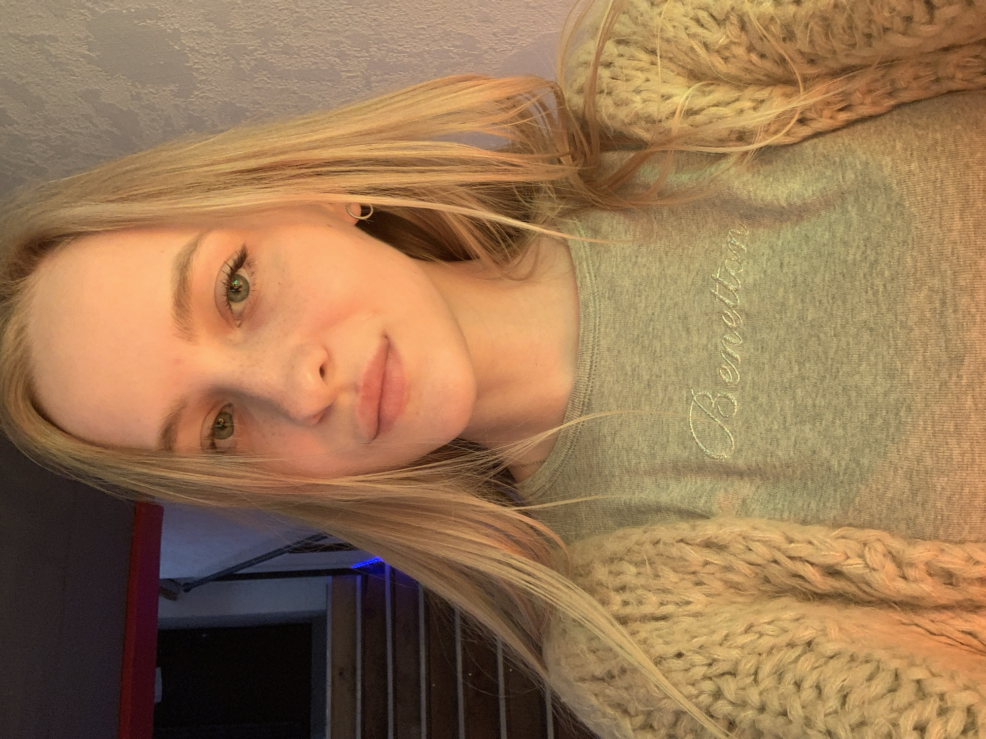 A young woman with blonde hair and light green eyes is looking at the camera. She is wearing a light gray long-sleeved shirt with "Benetton" embroidered in white on the chest, and a chunky knit cream cardigan. She has a small hoop earring in her right ear. Behind her, a staircase with wooden steps is visible, with blue and red lighting in the background. A textured wall is to her right. The scene appears to be indoors, possibly in a building with multiple levels or a basement area, with the lighting suggesting it might be evening or a dimly lit space. The location is the city of Kyiv, Ukraine.