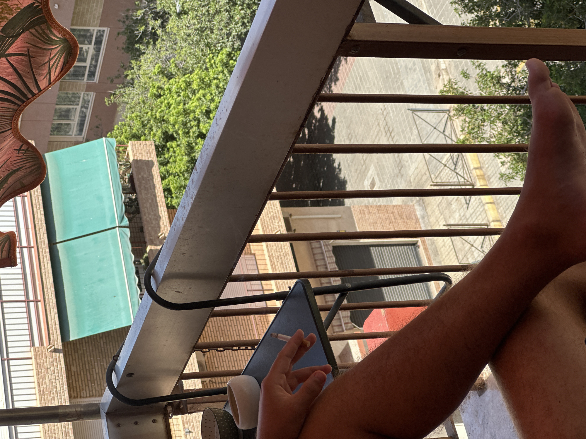 A medium shot captures a person's bare leg and foot resting on a balcony. The person's right leg, tanned and hairy, is crossed over their left leg, which is out of frame. A foot with visible toes is positioned in the lower right corner of the image. The person is holding a lit cigarette in their right hand, which is resting on a small black table. A white ashtray and a patterned bowl are also on the table. The balcony has a metal railing and a concrete floor. In the background, a building with brick accents and green awnings is visible, along with lush green trees and a paved area. The scene appears to be set in an urban environment.