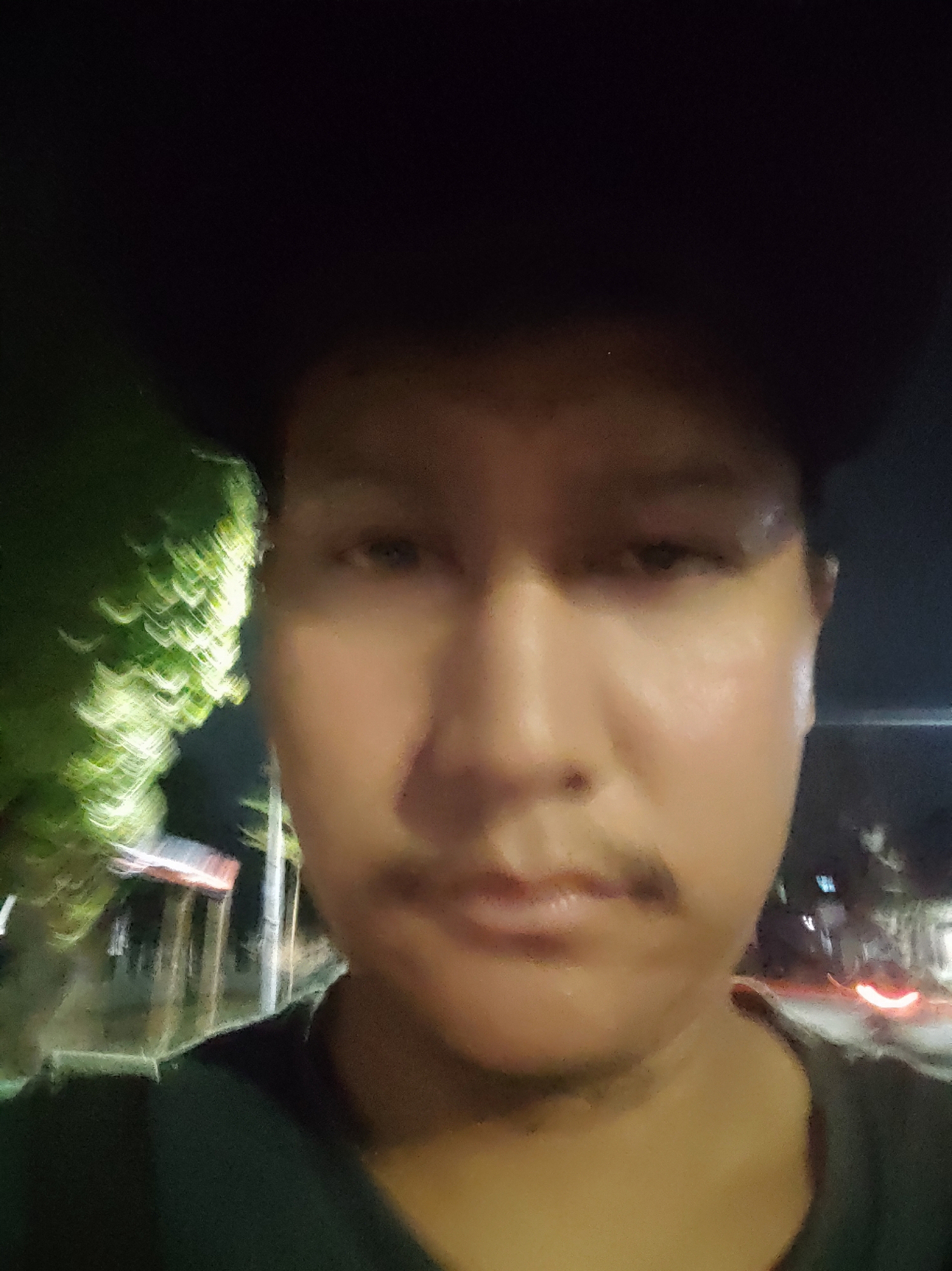 The image is a close-up selfie of a person's face taken at night. The person is wearing a dark hat that covers their forehead. The background is blurred and shows a street scene with some lights and foliage, suggesting an outdoor urban environment in Babakangebang, Indonesia. The lighting is dim, and the image quality is slightly grainy, typical of night photography.