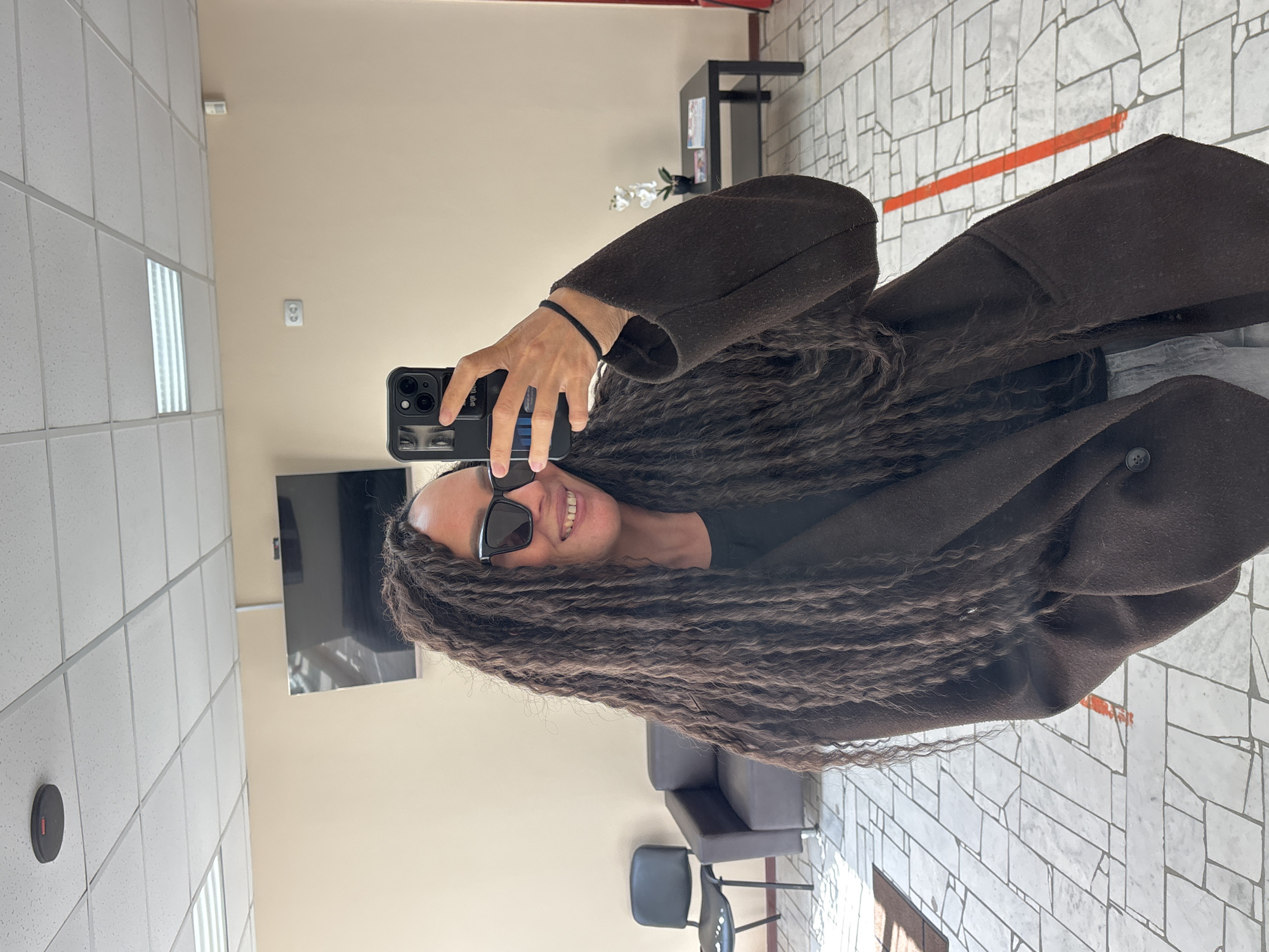 A woman with long brown braided hair is taking a selfie in what appears to be a public space. She is wearing sunglasses and a dark brown coat. The background includes a light beige wall, a black television mounted on the wall, a black shelving unit, seating, and a tiled floor with an orange painted stripe. Based on the location specified, this image was taken in Chaikovsky, Russia.