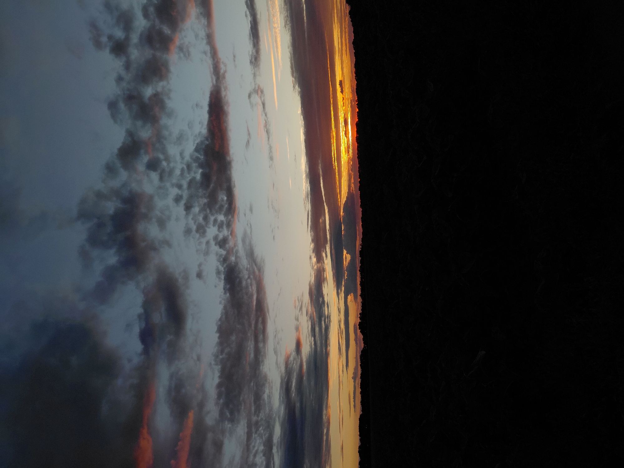 A dark silhouette of a treeline divides the frame horizontally. Above the treeline, the sky displays a dramatic sunset with a gradient of colors. Dark, textured clouds are scattered across the upper portion of the sky, tinged with hints of pink and orange where they catch the light. Lower in the sky, a bright band of orange and yellow illuminates the horizon, indicating the setting sun is just below the visible edge of the earth. The foreground consists of a dark, textured expanse, suggesting a field or open land under the twilight sky. The overall impression is one of a natural landscape at dusk. The location is described as Unknown, Unknown.