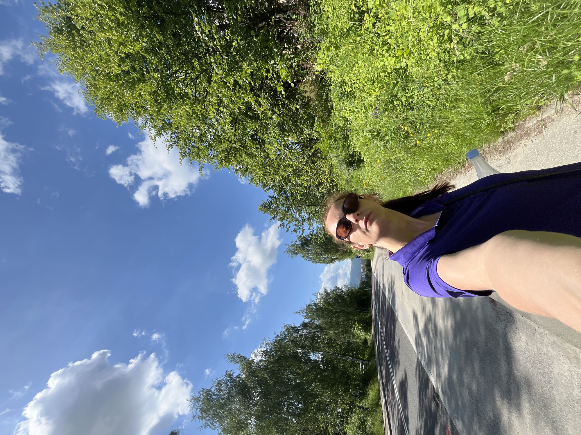 A woman wearing sunglasses and a purple polo shirt takes a selfie outdoors. She is standing on a sidewalk next to a road, with trees and lush green vegetation lining the right side of the road. The sky above is blue with scattered white clouds, and the sun casts shadows on the road and sidewalk. The woman is holding a clear plastic bottle with a blue cap in her right hand. The road appears to be a two-lane road with white markings. The background shows more trees and distant buildings. The scene suggests a pleasant day outdoors, possibly during a walk or hike.