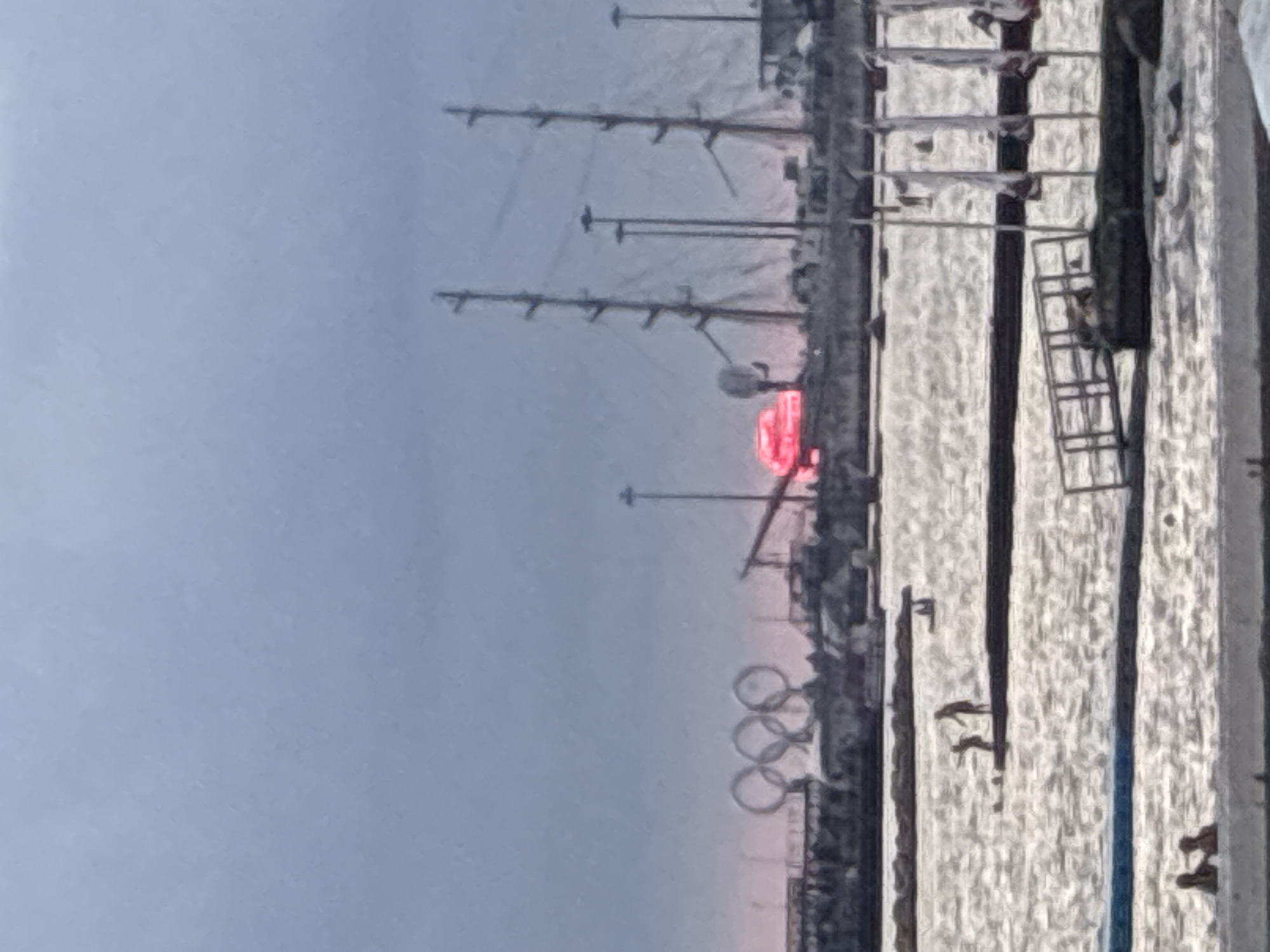 The image depicts a waterfront scene in Sochi, Russia, likely at sunset. In the background, the Olympic Rings symbol is visible against a hazy sky. Behind the rings, a silhouette of buildings and structures can be seen, including what appears to be a ship with multiple masts. A bright red, circular object, possibly the setting sun or a light, is positioned to the right of the Olympic Rings.
In the mid-ground, the water is choppy and reflects the subdued light. Several figures are present on a floating platform or walkway. Two individuals are standing, one appearing to point or gesture towards another, while a third person is partially visible in the water. Further into the water, another individual is seen swimming or wading.
The foreground of the image shows the edge of a pool or a similar water feature with two figures partially submerged. A metal ramp leads into the water from a dark platform. The overall impression is that of a recreational area by the sea, with elements of the city's Olympic past visible in the background. The sky is overcast, with a muted color palette dominating the scene.