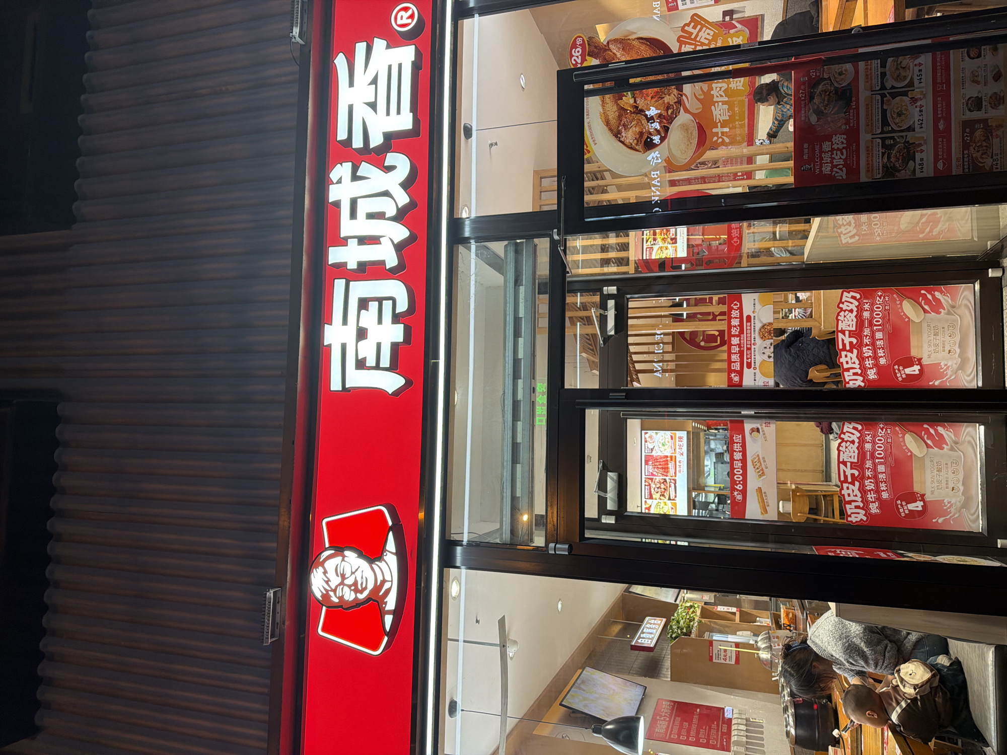 The image captures the storefront of a Nan Cheng Xiang restaurant, a popular fast-food chain in China, likely located in the Dongcheng District based on its prevalence in Beijing. The restaurant's bright red signage prominently displays the brand name and logo. The store is at night. A woman can be seen through the window, attending to a child. Inside, the restaurant's interior is visible, featuring tables and chairs. The entrance is characterized by large glass doors and windows, adorned with promotional posters for various food and beverage items, including milk skin yogurt. The street in front of the restaurant has a metal door on the side of the restaurant.