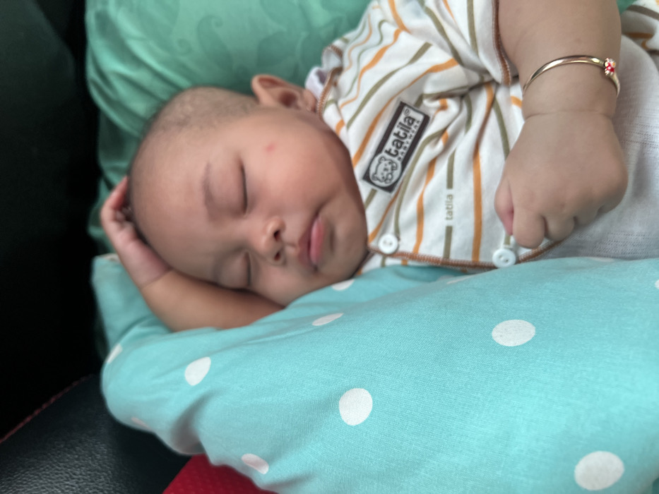 A close-up image depicts an infant, likely of Asian descent, sleeping. The infant's eyes are closed, and its mouth is slightly open. Its left hand rests on the top of its head, and a faint red mark is visible on its left cheek. The infant is clad in a light-colored, striped onesie featuring narrow orange and beige horizontal stripes, secured with white snap buttons along the front. A dark rectangular label on the chest of the onesie displays "TATILA BABYWEAR" and a bear graphic. A thin, gold-toned bracelet with a small red, multi-petal charm is present on the infant's right wrist. The infant is positioned on a teal-colored, wrinkled soft surface, likely a pillow or blanket. To the left of the infant, a blue textile with distinct white polka dots is visible. In the upper left background, a dark, padded surface with red stitching, consistent with vehicle interior upholstery, is present. The precise location within Tasikagung, Indonesia, cannot be determined from the visual data.