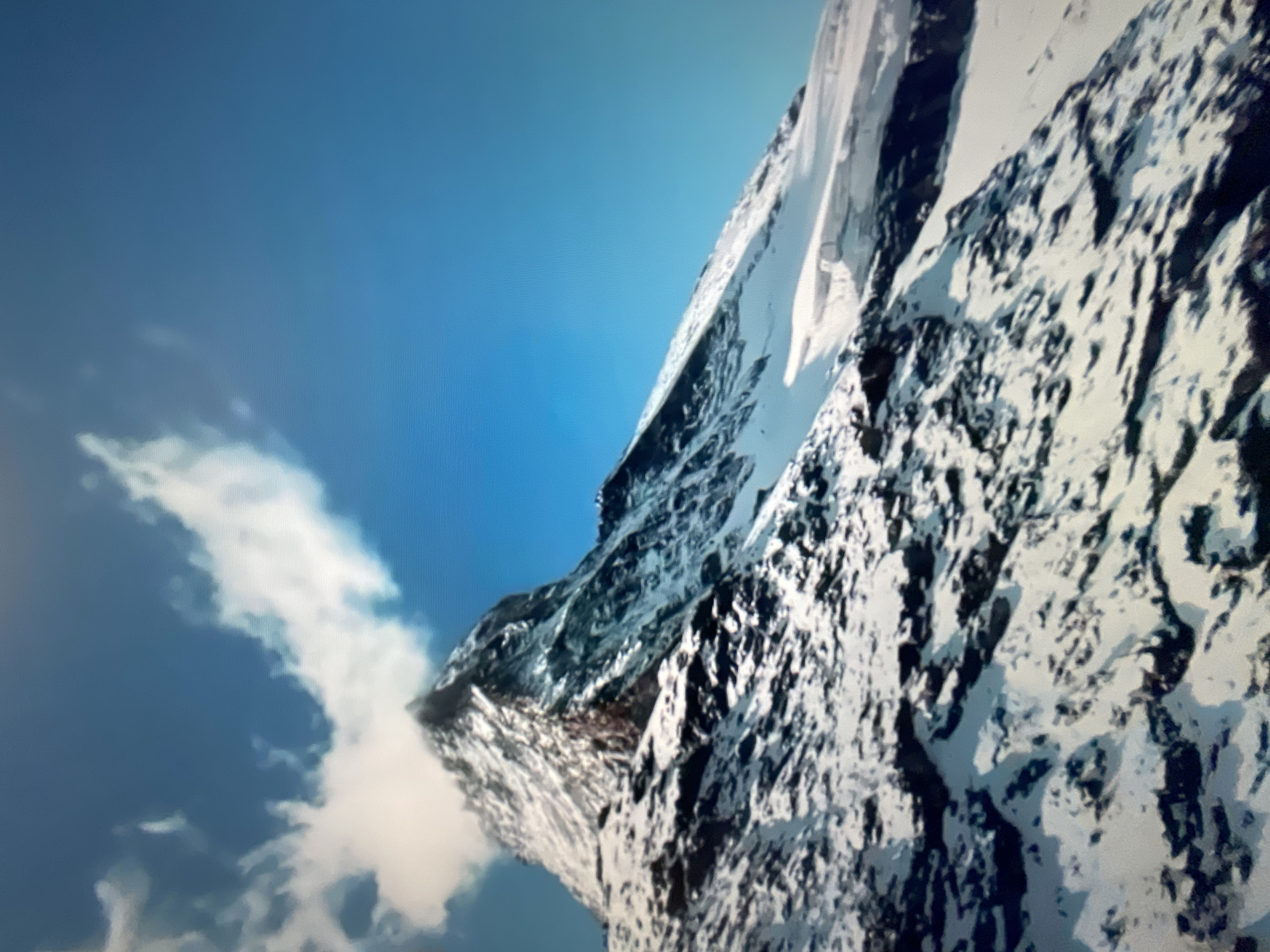The image depicts a snow-covered mountain peak under a bright blue sky with scattered white clouds. The mountain is rocky and rugged, with patches of snow clinging to its slopes and summit. The lower portion of the mountain is more densely covered in snow, interspersed with dark, rocky outcrops. A large cloud hovers near the summit, partially obscuring it. The scene is a natural landscape, devoid of any urban elements or human presence. There is no indication of the city of Kafr Yasif, Israel, in this image.