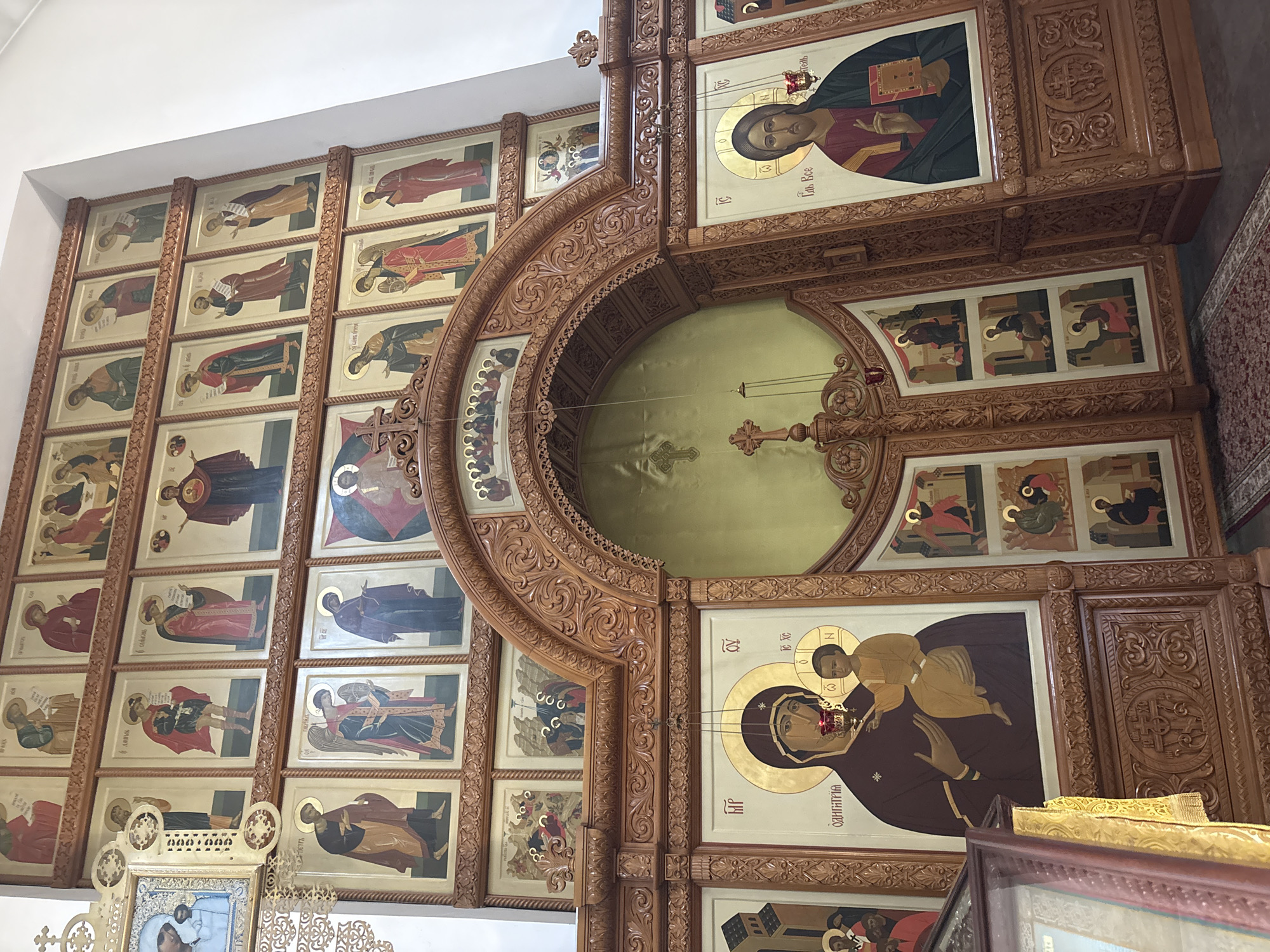 The image depicts the interior of the Church of the Iberian Mother of God in Sestroretsk, Russia. The focal point is the ornately carved wooden iconostasis, which dominates the scene. The iconostasis features multiple rows of religious icons, painted in a traditional style, depicting various saints and biblical scenes. The icons are framed by intricate wooden carvings. In the center of the iconostasis, a large arched opening reveals a gilded backdrop. Flanking this opening are two large icons; one depicts the Virgin Mary holding the Christ child, and the other shows Jesus Christ. The wooden structure extends across the frame, with various carved panels and decorative elements. The overall impression is one of a richly decorated and spiritual environment within a church.