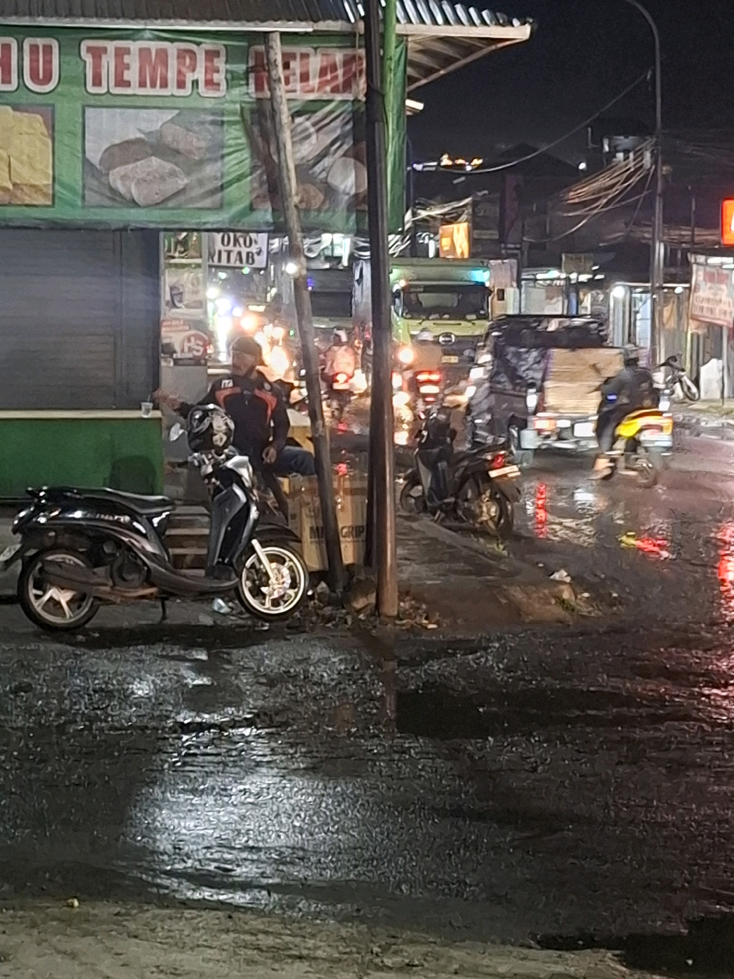 The image depicts a nighttime scene on a street in the city of Ciheulang Tonggoh, Indonesia. A man in a dark jacket with orange trim is seated on a black scooter. The scooter is parked on a wet, dark asphalt surface, reflecting the surrounding lights. To the left of the scooter, there is a green awning with text in red and white, suggesting a food stall or shop, with an image of tempeh on display. Behind the scooter, a truck is visible, its cargo covered. Further in the background, there are other vehicles and a traffic light. The overall atmosphere is urban and bustling, with wet conditions indicating recent rain.