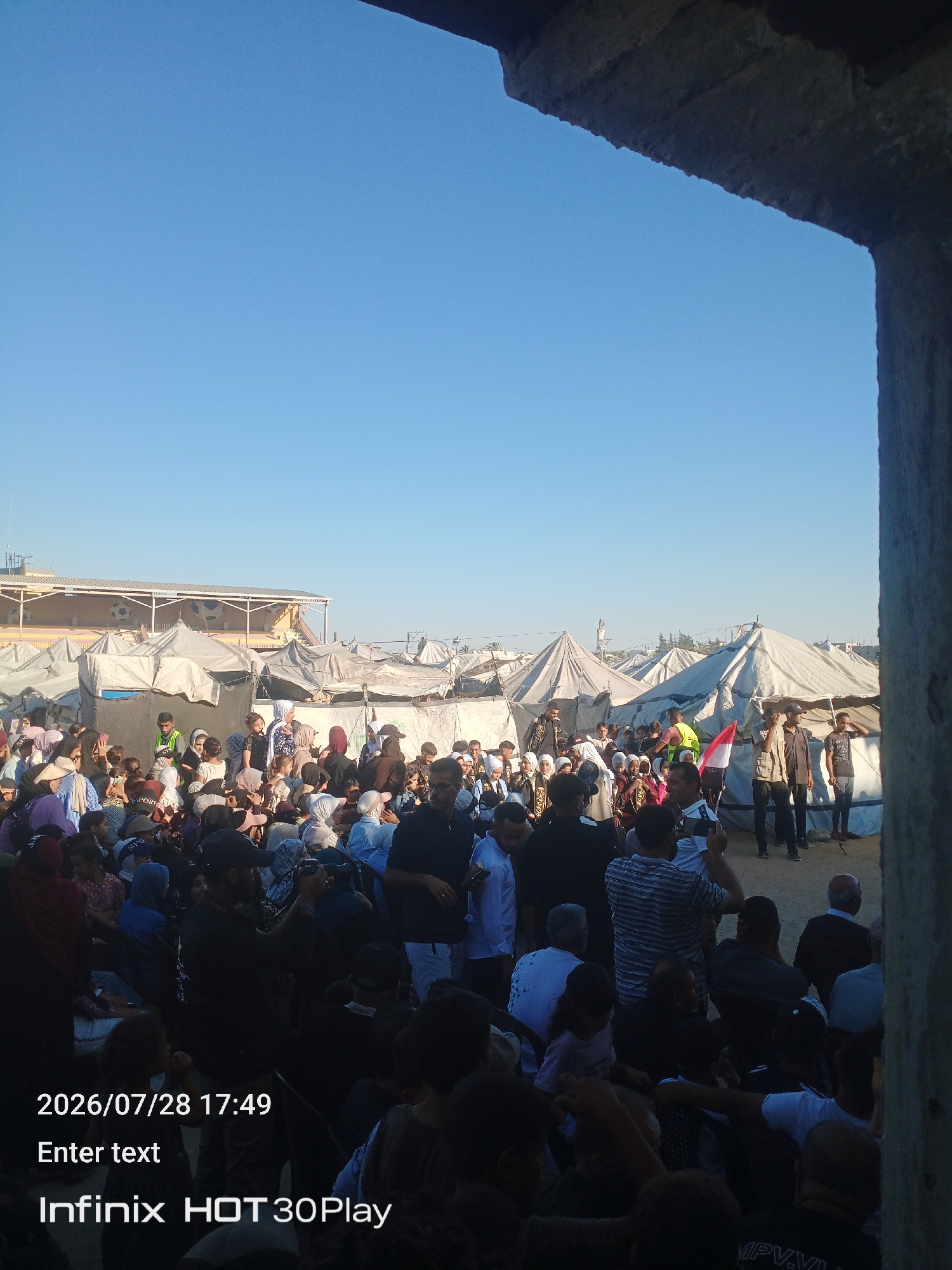 This image depicts a crowded outdoor gathering of people amidst tents, likely a refugee or displacement camp, in the city of Nuseirat, Palestinian Territories. The sky is clear and blue. In the foreground, a concrete structure partially frames the scene. A large group of individuals, including many children and women, are visible. Some are standing, while others are seated. Tents of various sizes and colors, characteristic of temporary shelters, are set up behind the crowd. Several individuals appear to be taking photographs or videos with their phones. A red, white, and black flag, possibly a national or organizational flag, is being held up by a person in the middle ground. The overall atmosphere suggests a communal event or assembly within the camp. The bottom of the image contains overlaid text indicating the date and time "2026/07/28 17:49", the prompt "Enter text", and the device model "Infinix HOT30Play".
