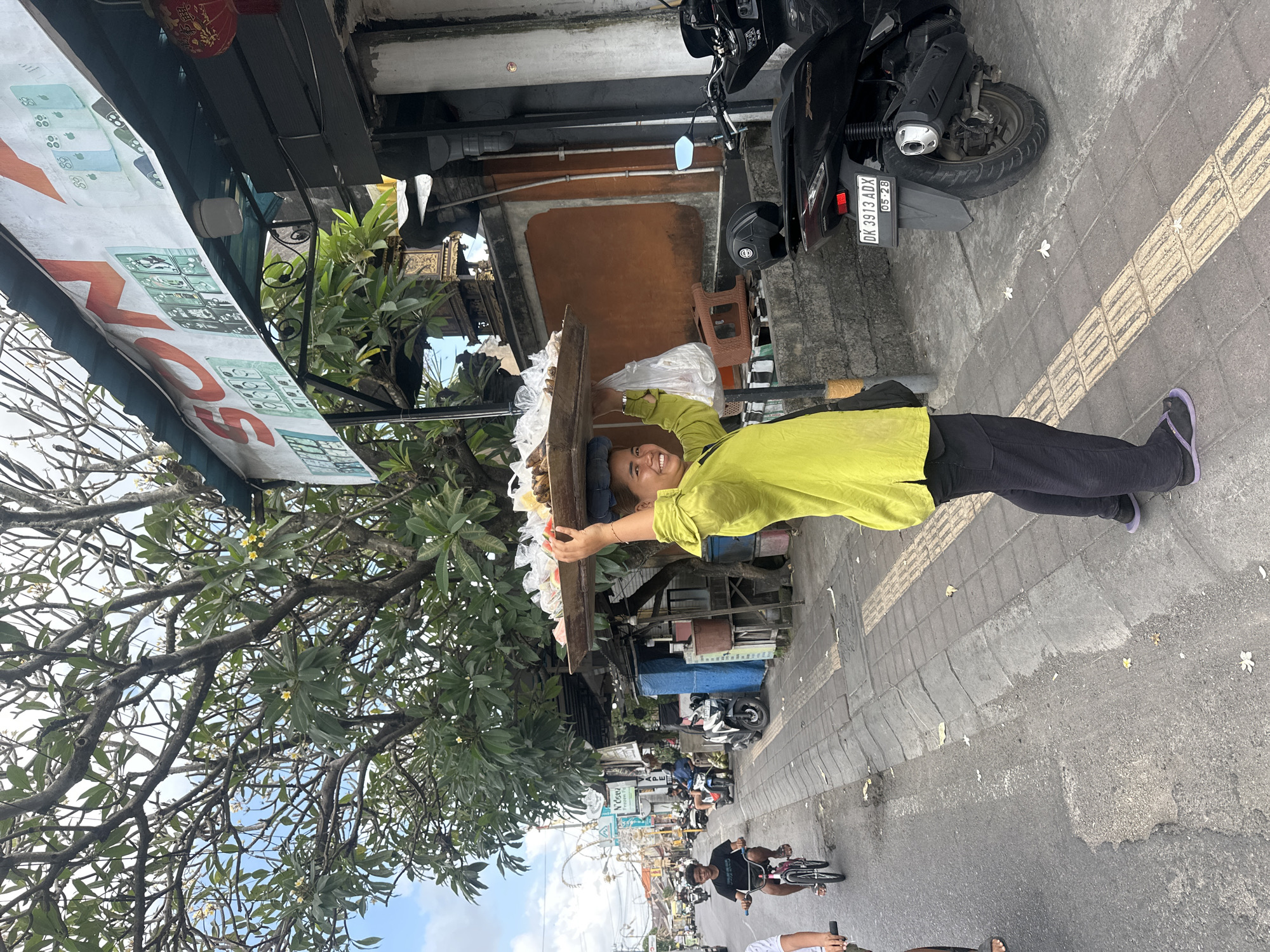 A woman in a bright lime green shirt and black pants, wearing flip-flops, balances a wooden tray of food on her head. She is smiling at the camera. The tray contains various items wrapped in plastic. A white plastic bag hangs from the tray. She is standing on a paved sidewalk next to a street. In the background, there is a street with several motorbikes and a young boy riding a bicycle. A scooter is parked on the sidewalk to her right. The street is lined with buildings and trees. The scene appears to be in Denpasar, Indonesia, based on the license plate of the parked scooter which reads "DK 3913 ADX" where "DK" is the registration code for Bali. The sign above the building in the background also appears to be in Indonesian.