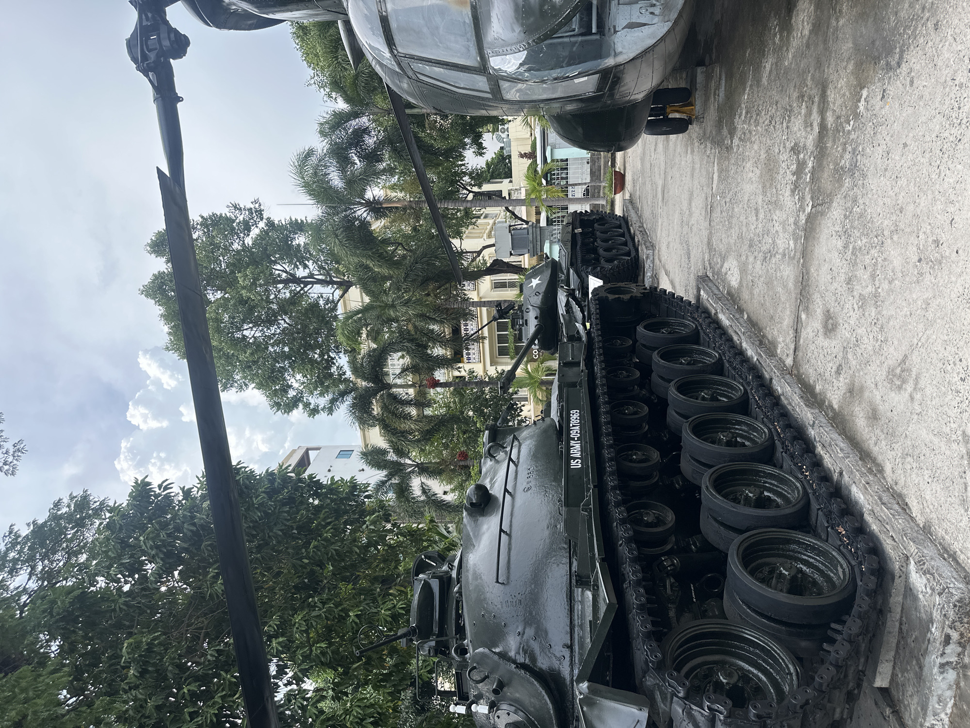 The image shows an outdoor display of military vehicles in what appears to be the city of Thủ Đức, Vietnam. A dark green tank with black treads and road wheels is positioned in the foreground. The tank has the inscription "US ARMY-09A78969" along its side. Behind the tank is a dark green military helicopter. The background includes palm trees, other trees, and buildings. The ground is concrete. The sky is overcast.