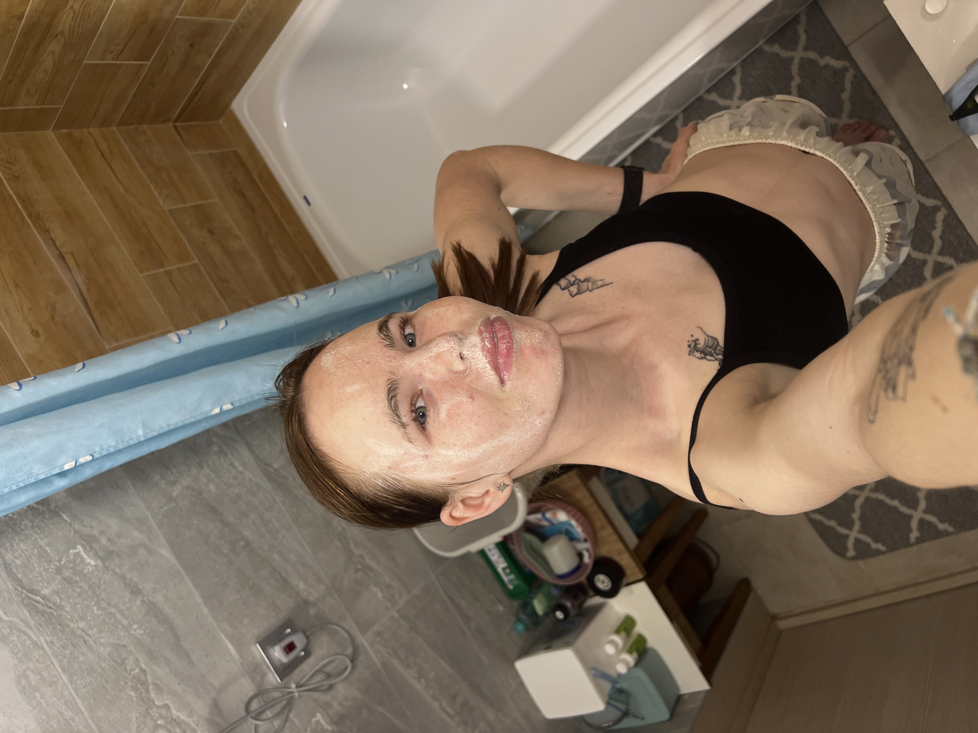 A selfie-style photograph, taken at an acute angle, shows a person in a bathroom. The person is wearing a black top and a shower cap, with what appears to be a facial mask applied. Tattoos are visible on their chest and arm. The bathroom features a bathtub with a blue shower curtain, gray tiled floors, and wooden-paneled walls. Various toiletries and cleaning products are arranged on a small white cabinet. No location in Perm, Russia, is identifiable within the image.
