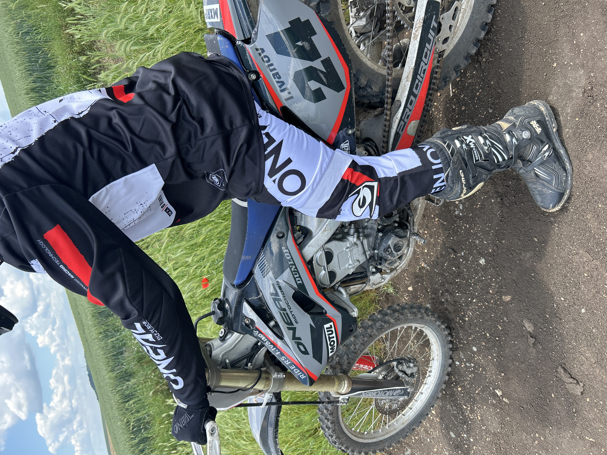 The image captures a person riding a dirt bike on what appears to be a dirt path alongside a grassy field. The rider is wearing a black and white O'Neal brand racing suit with a red accent, O'Neal gloves, and black Forma XW Series motocross boots. The dirt bike has O'Neal and Dunlop branding, with the number "24" and the name "I. Ivanov" printed on the side. The motorcycle is positioned on a dirt path, with a field visible in the background under a bright, partly cloudy sky.