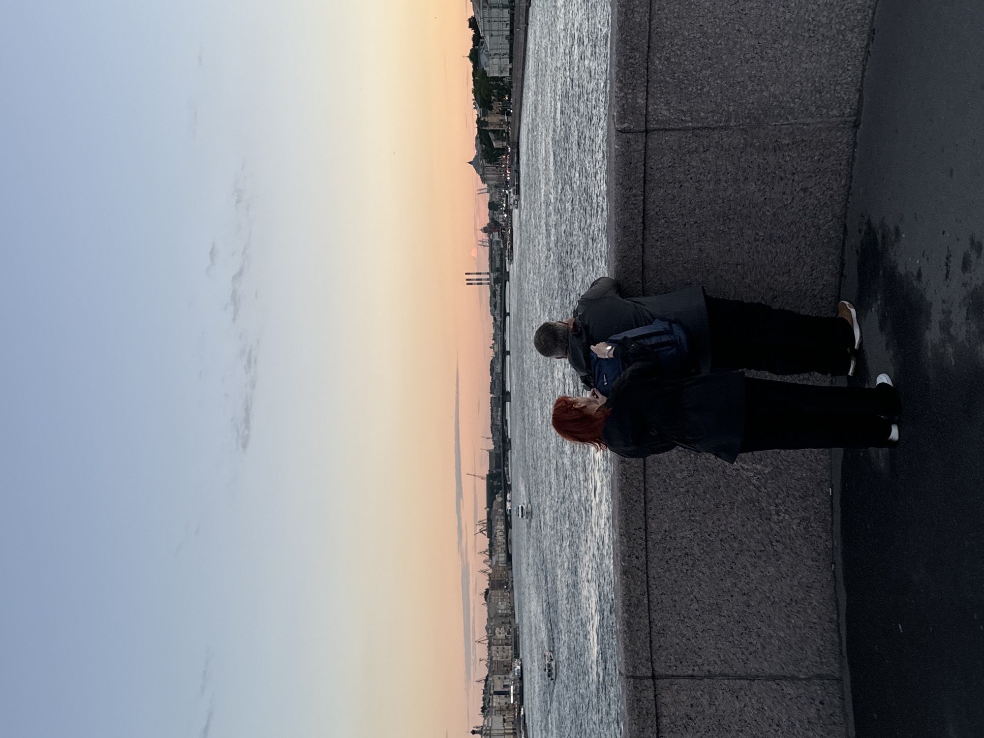 This is a vertical, eye-level, full-body shot of two people standing on a stone embankment overlooking a wide river in St. Petersburg, Russia, at dusk. The sky is a gradient of soft blues and oranges, indicating the sun has recently set or is about to rise. In the distance, the city skyline is visible, featuring numerous buildings, bridges, and industrial cranes. Several boats are on the river, including two small white passenger vessels.
In the foreground, a woman with bright red hair stands on the left, facing away from the camera and looking towards the river. She is wearing a dark jacket and black pants, with white sneakers. Next to her, on the right, a man also stands facing away, his back to the viewer. He is wearing a dark hooded jacket and black pants, with brown sneakers. He has a blue backpack slung over his left shoulder. The two individuals are standing close together, suggesting they are a couple or companions. The stone embankment they are standing on is textured and gray, with a dark asphalt surface in front of it.
