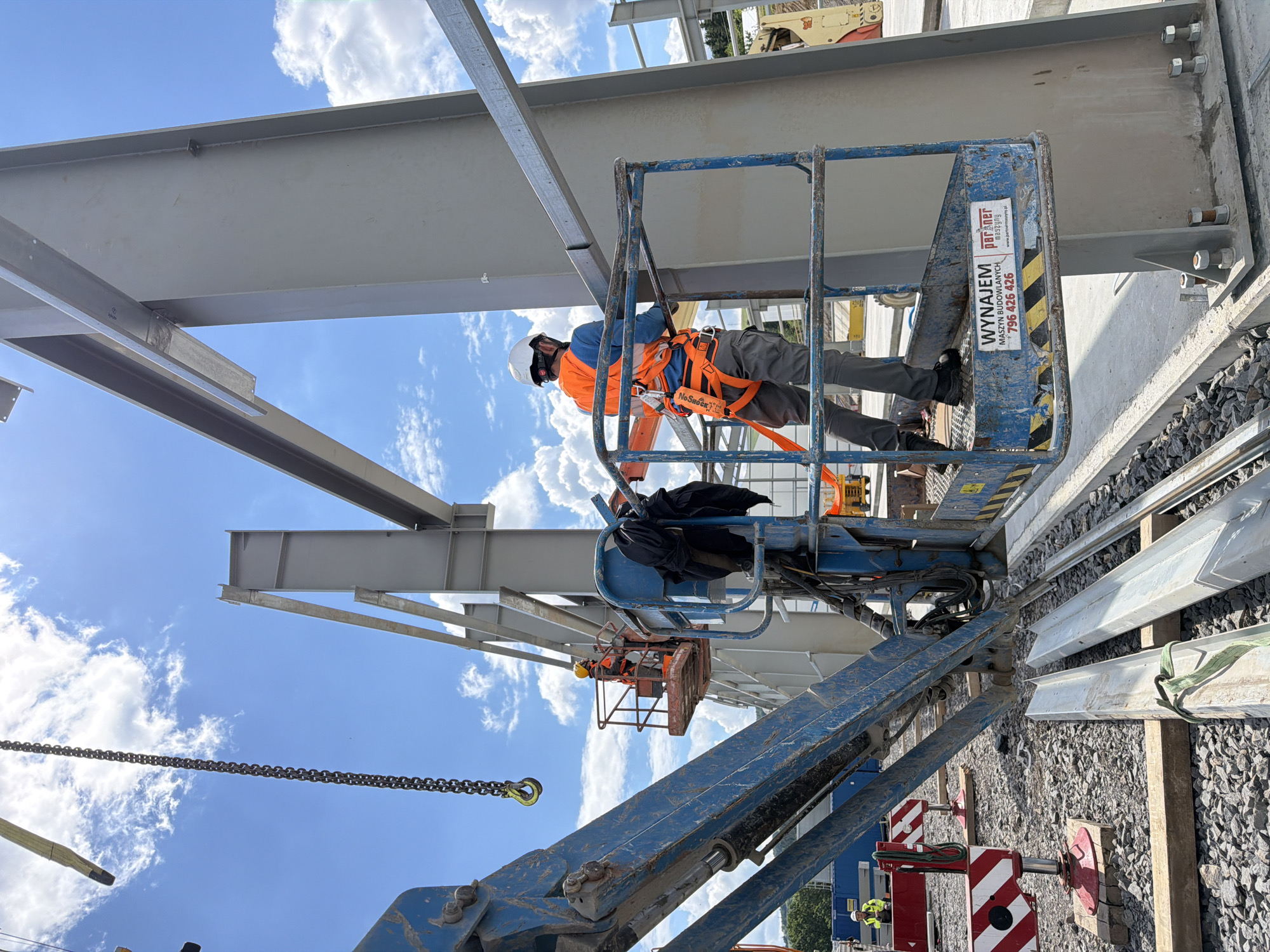A construction worker wearing a white hard hat, blue shirt, orange safety vest, and grey pants is standing in the basket of a blue aerial work platform. The worker is holding onto the railing of the basket, and appears to be inspecting or working on a large steel beam that is part of a larger structure under construction. Another worker in a yellow hard hat and orange vest is visible in the background, also in an aerial work platform. The scene is taking place outdoors on a sunny day with a blue sky and white clouds. The ground is covered with gravel and construction debris. Several pieces of steel construction material are lying on the ground. A large crane chain hangs from above and to the left of the frame. A sign with Polish text and a phone number is affixed to the side of the aerial work platform. The location is identified as Janowice, Poland.