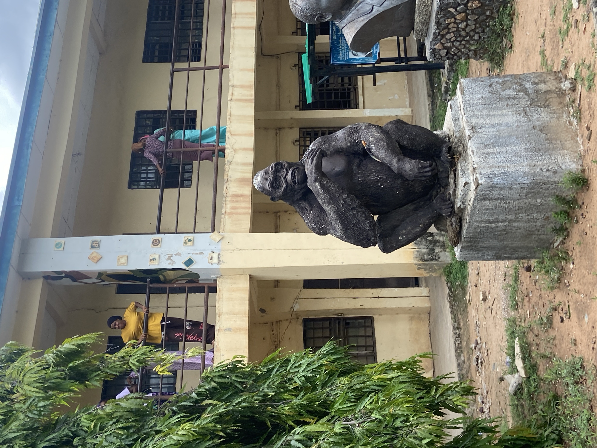 The image shows a statue of a primate, potentially a gorilla, located on the grounds of a building in Lokoja, Nigeria. The statue is dark-colored and positioned on a weathered concrete base. Behind the statue is a multi-story building with barred windows. People are visible on the balconies of the building, with one person wearing a yellow shirt and another wearing a floral print top and blue pants. The foreground features a leafy green bush. The ground is a mix of dirt and sparse grass. Another statue, partially visible, is positioned behind the primate statue.
