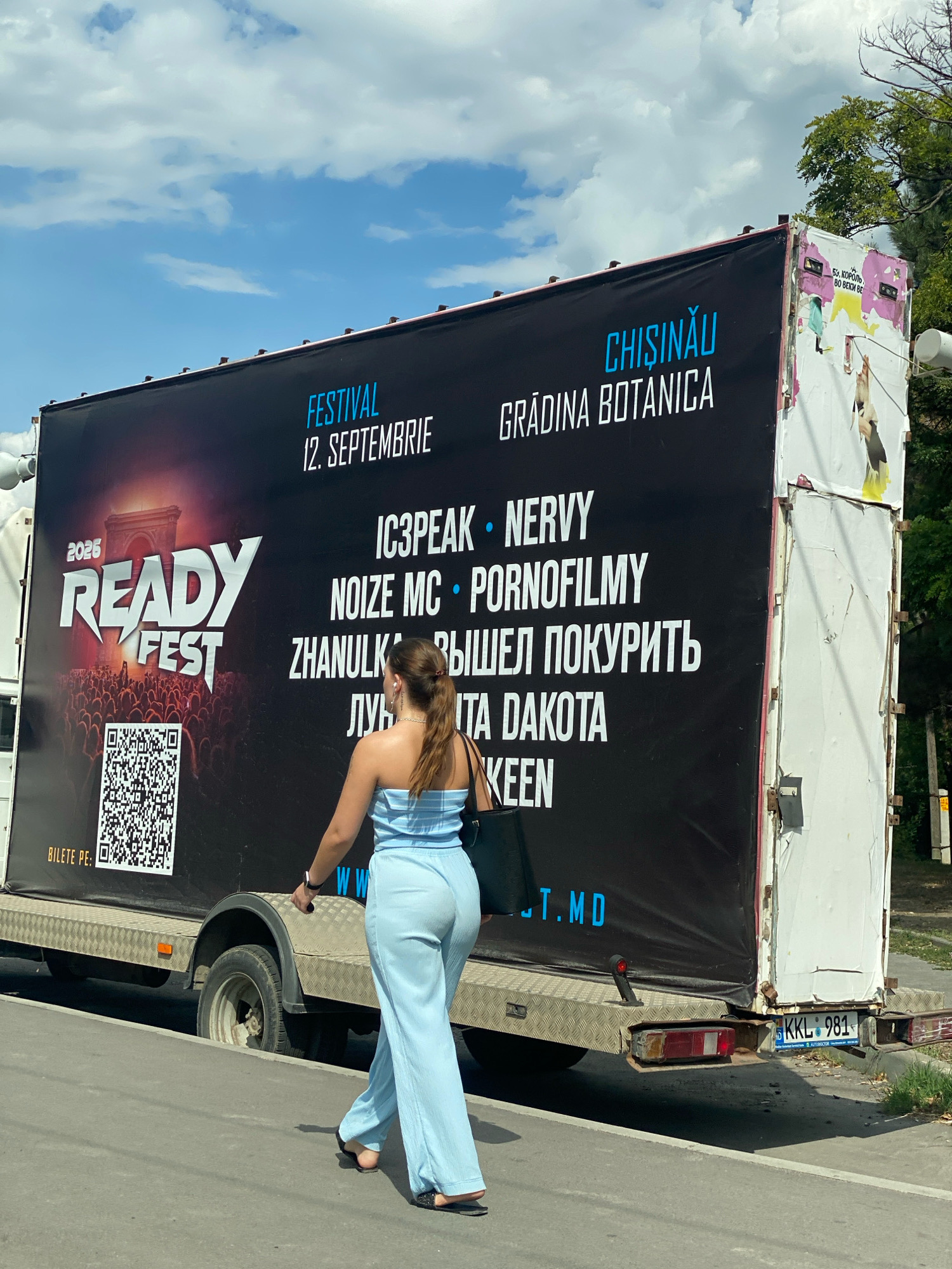 A low-angle, medium shot captures a large, black banner advertising a festival, attached to the side of a trailer. The banner reads "2025 READY FEST" in large white and red lettering on the left, with a crowd silhouetted in front of a monument. To the right, in blue and white text, are details for "FESTIVAL 12. SEPTEMBRIE", "CHIȘINĂU GRĂDINA BOTANICĂ", and a list of performers including "IC3PEAK • NERVVY", "NOIZE MC • PORNOFILMY", "ZHANULK • ВЫШЕЛ ПОКУРИТЬ", "ЛУЧШИТА DAKOTA", and "KEEN". Below the performer list, the website "W.W. .T.MD" is visible. To the far left of the banner is a QR code and the text "BILETE PE:". A woman, seen from behind and wearing a light blue strapless top and matching wide-leg pants, walks away from the viewer towards the right, her left hand holding a black tote bag. The trailer is parked on a paved surface, with grass and trees visible in the background. A license plate on the rear of the trailer reads "KKL 981". The sky above is blue with scattered white clouds.
